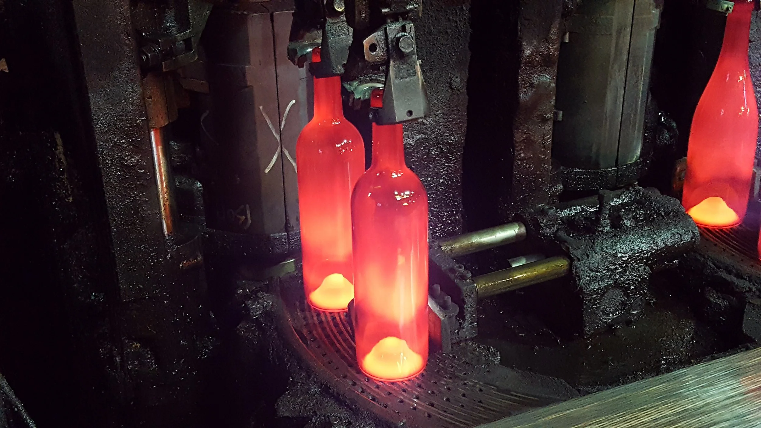 IS glass manufacturing process where glowing orange glass bottles are being shaped or cooled on a conveyor belt, with an industrial furnace in the background.