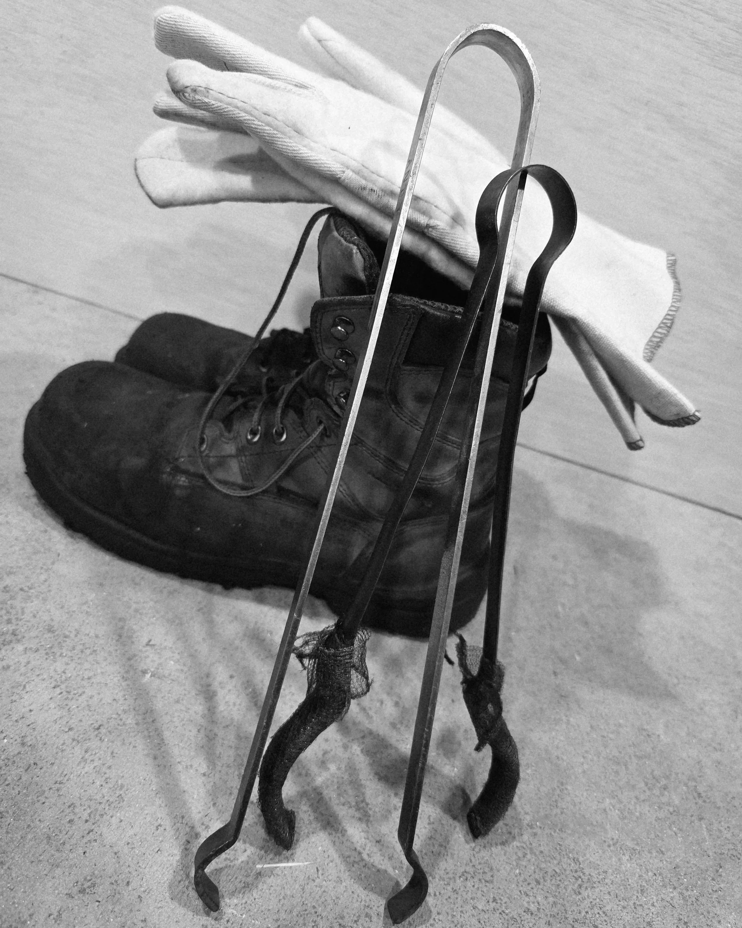 A worn black work boot, a pair of gloves, and a pair of large metal tongs are arranged on a wooden surface.
