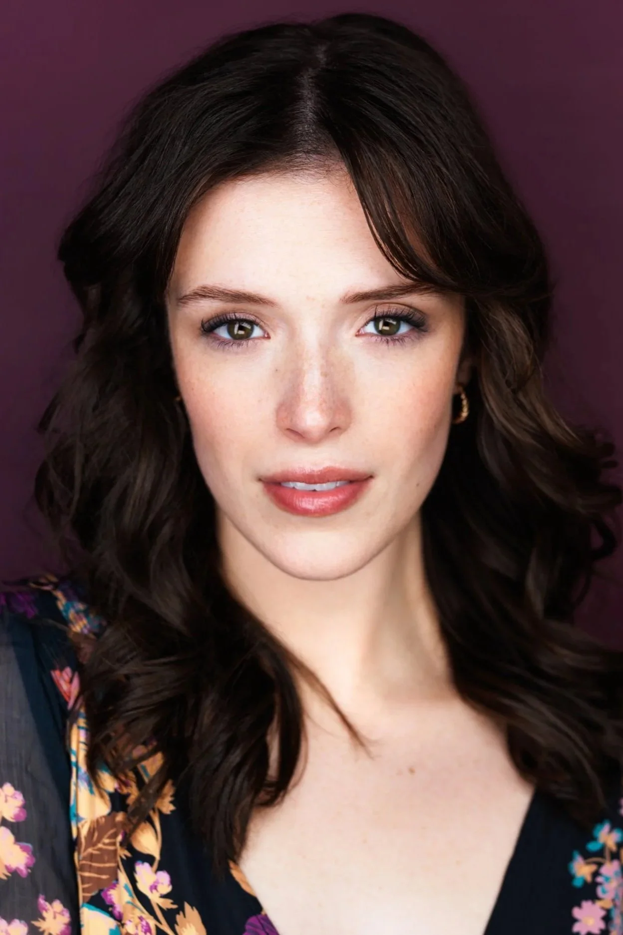 Close-up portrait of a young woman with wavy dark brown hair, light skin, hazel eyes, and a subtle smile, wearing earrings against a purple background.