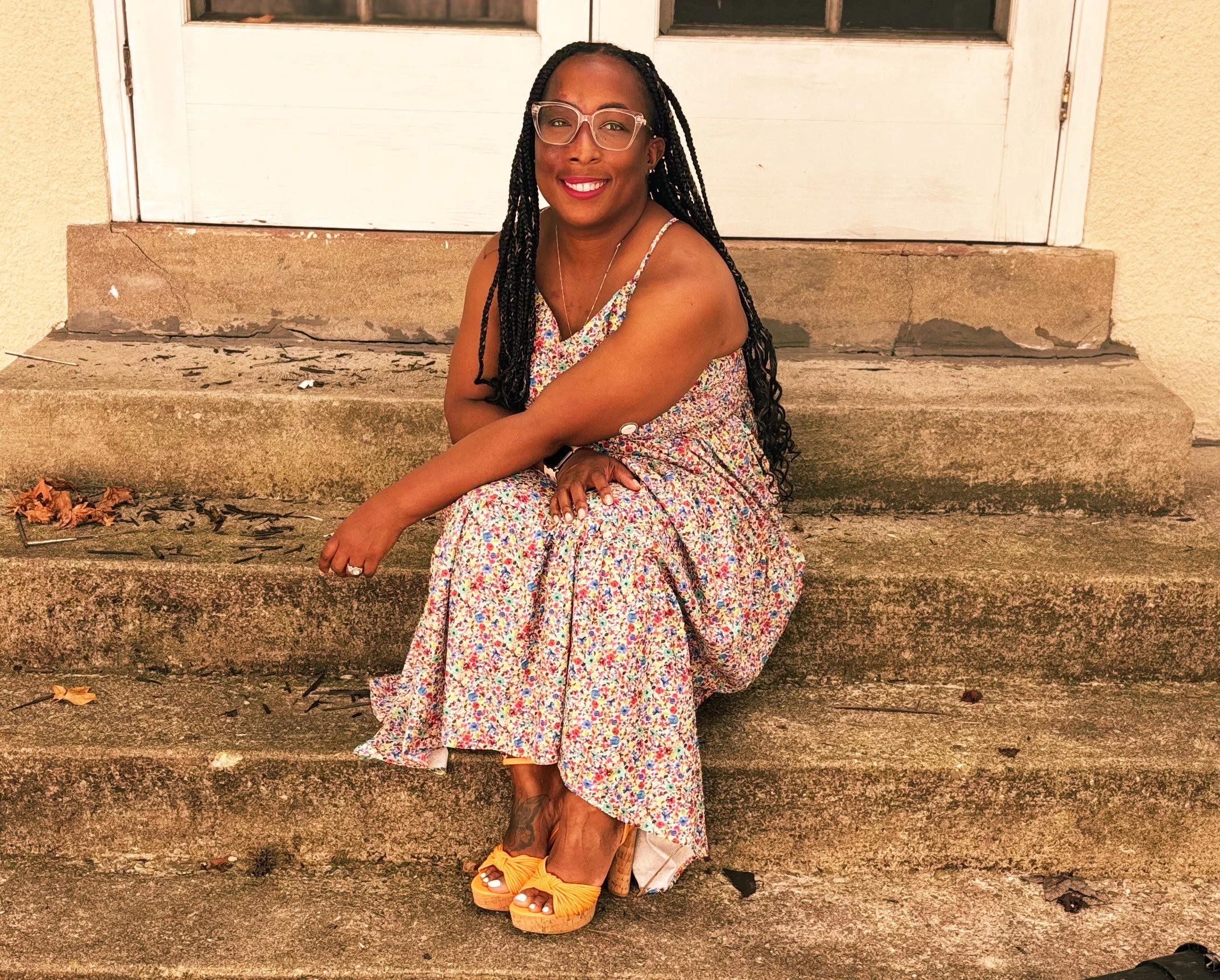 A woman with long braided hair, wearing glasses, a floral dress, and yellow sandals, sitting on concrete steps outside a door, smiling at the camera.