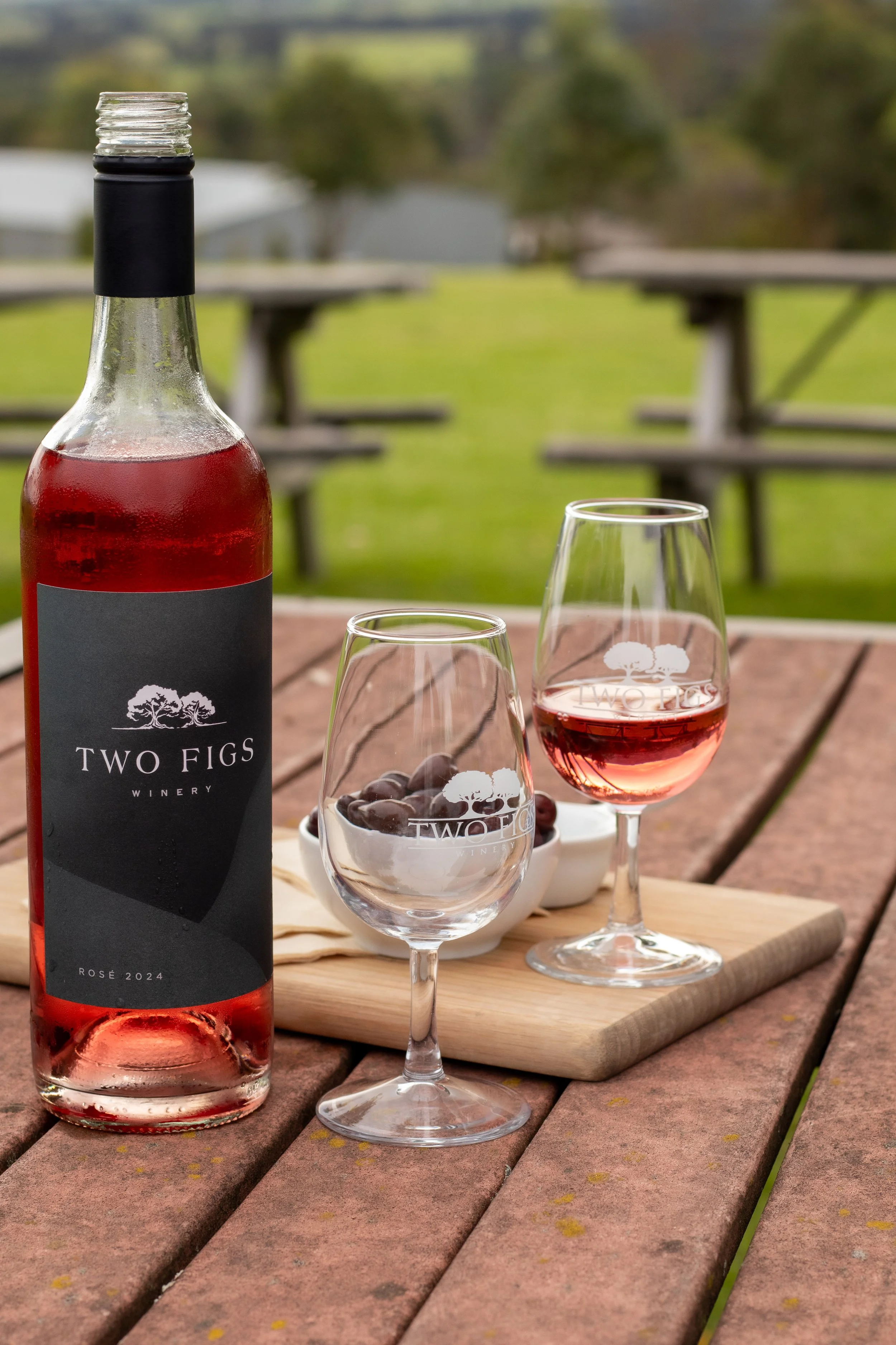A bottle of rose wine from Two Figs Winery, two wine glasses with pink wine, and a bowl of grapes on a wooden table outdoors.
