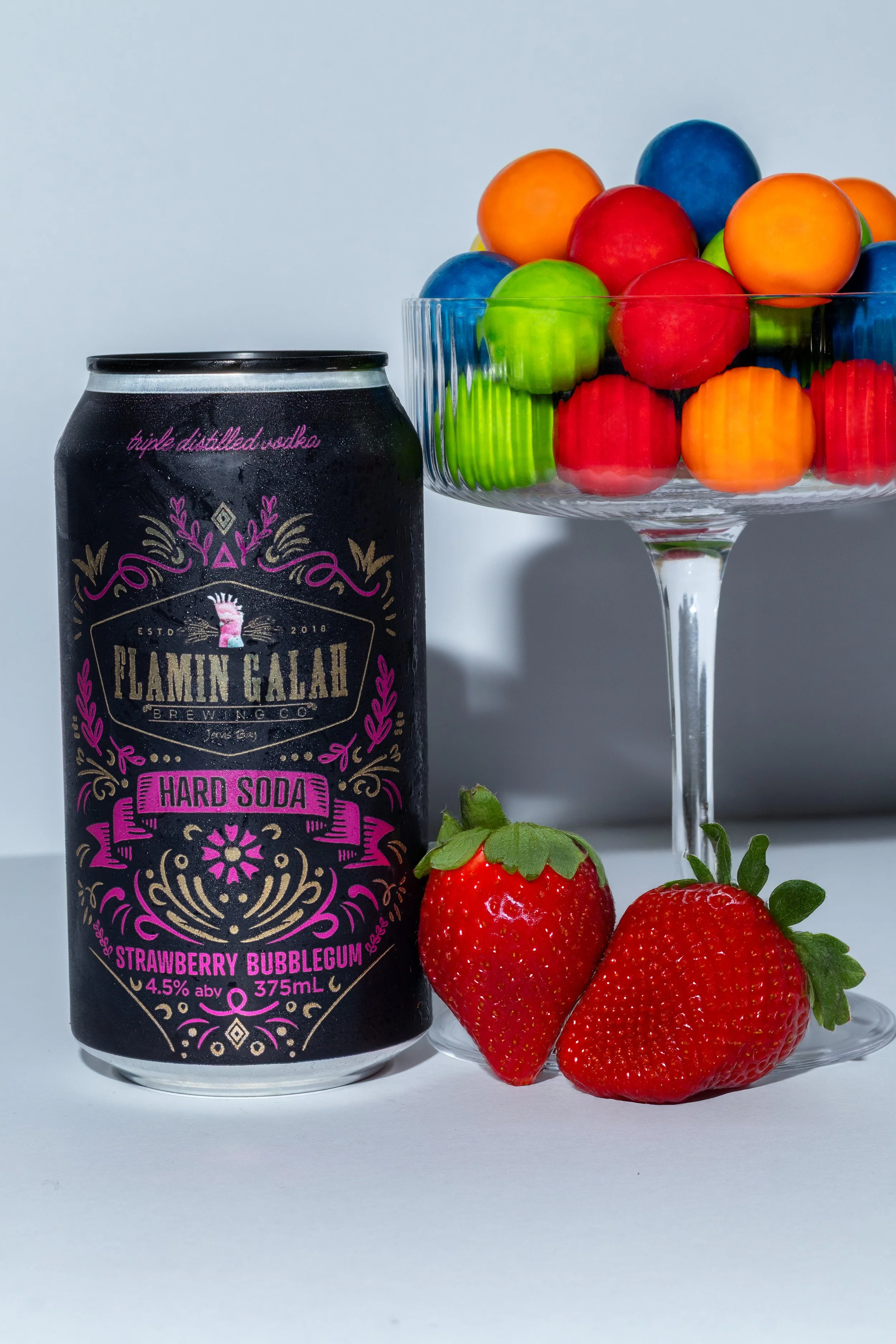 Can of Flamin' Gala Strawberry Bubblegum Hard Soda next to two strawberries, a glass bowl of colorful gumballs, and a white background.