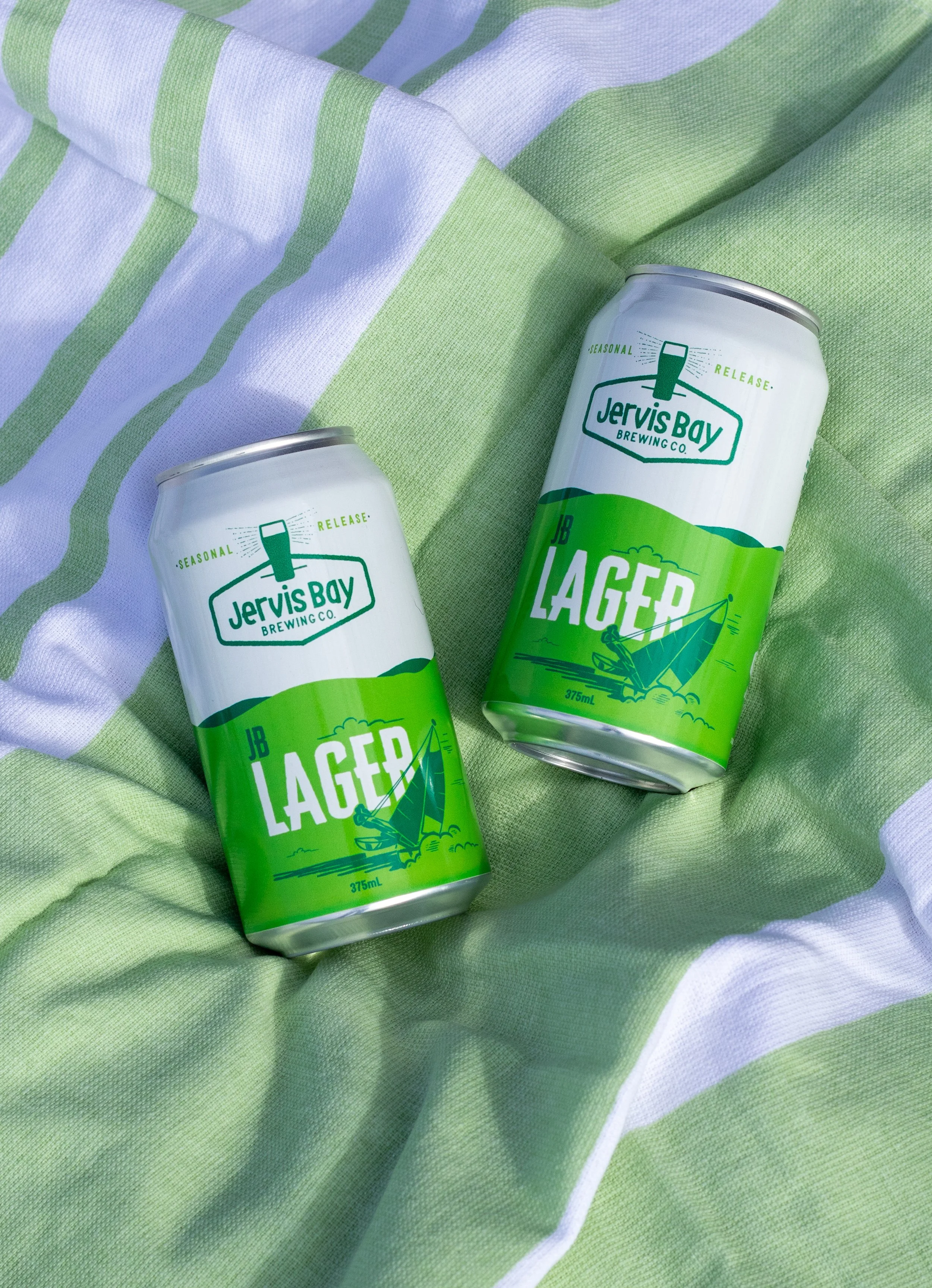 Two cans of JB Lager beer from Jervis Bay Brewing Co. placed on a green and white striped towel.
