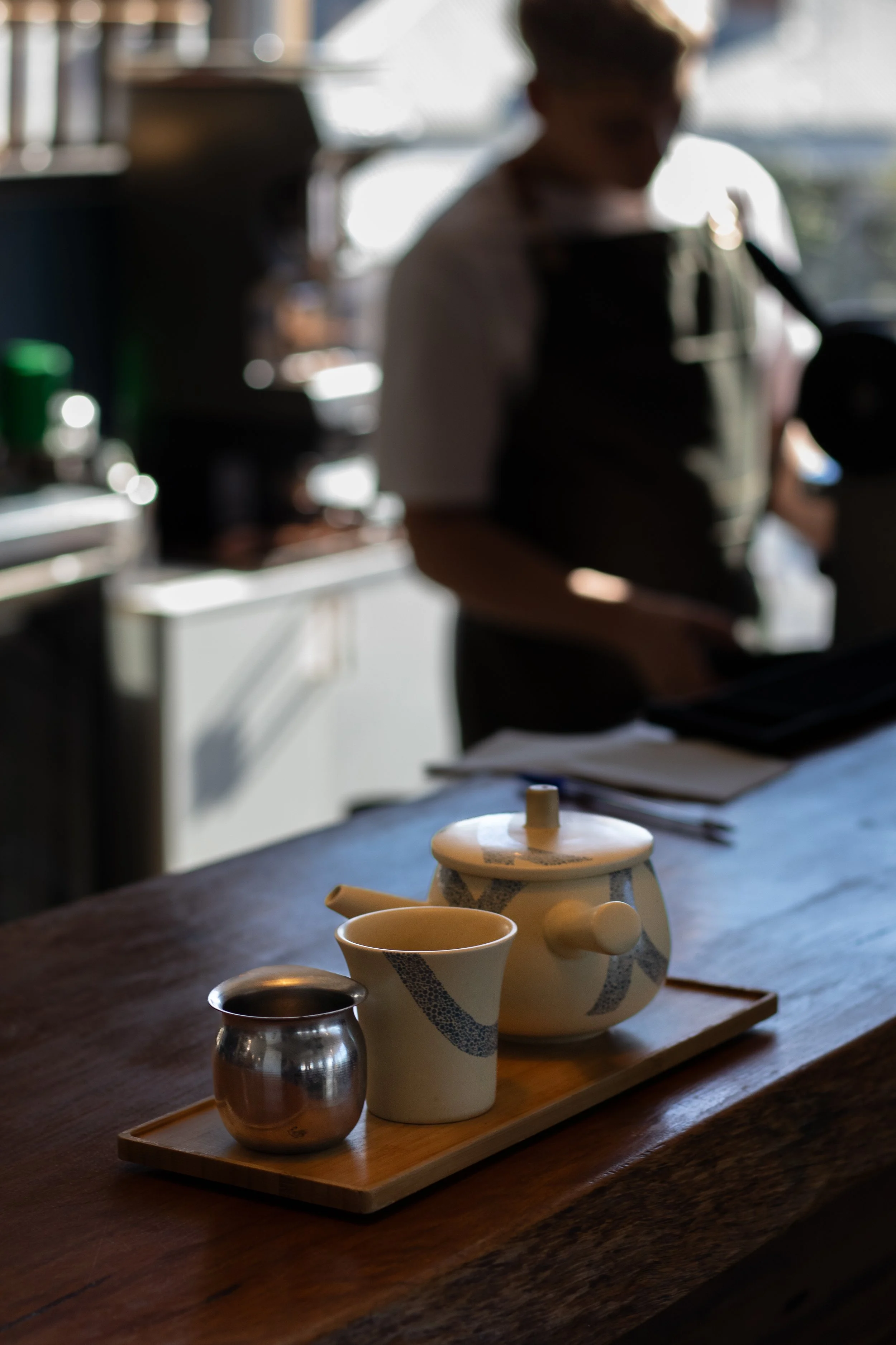 Tea set with a teapot, a small metallic pitcher, and a cup on a wooden tray on a table in a cafe. In the blurry background, a person is preparing drinks.