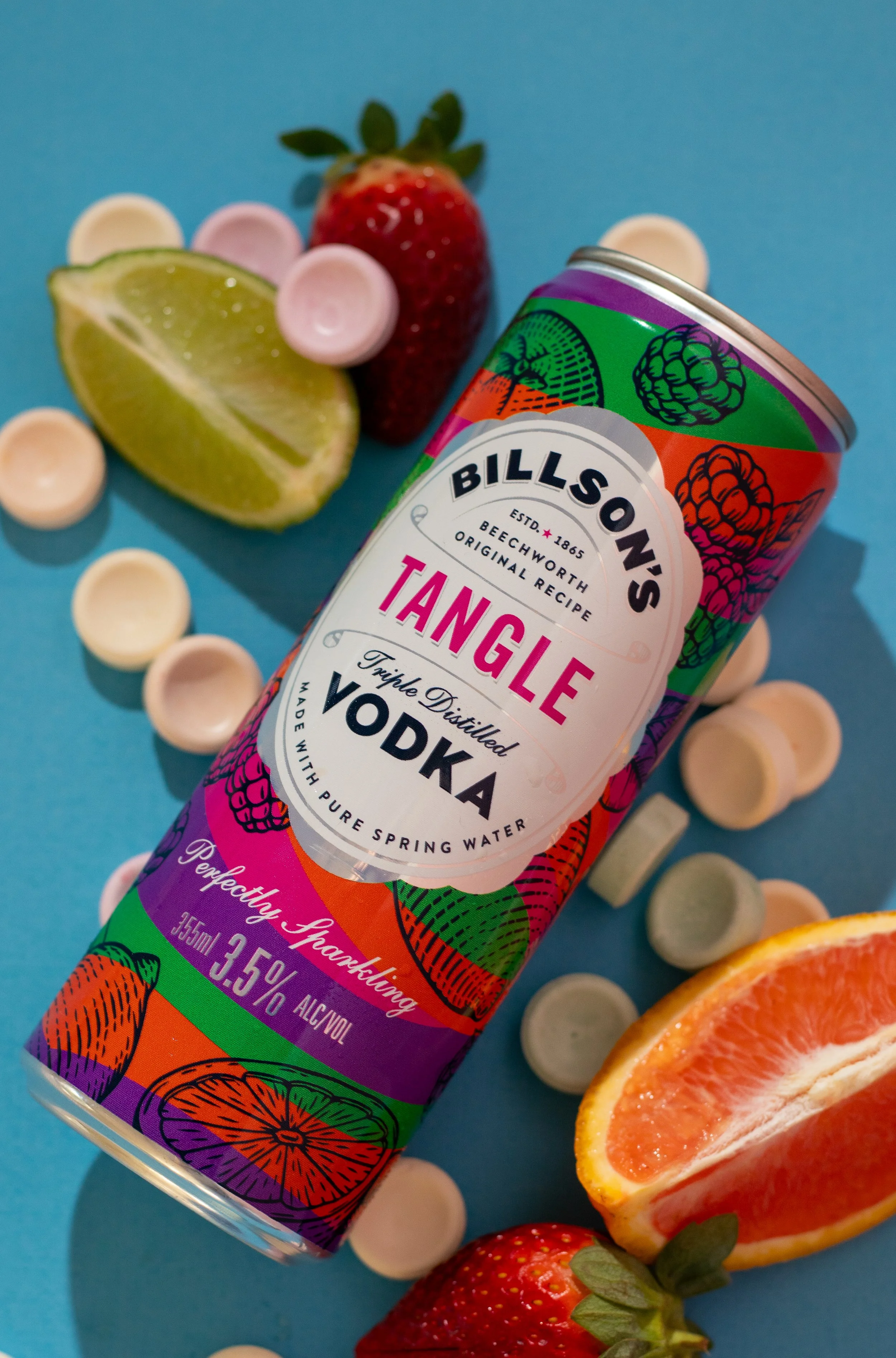 A colorful can of Billson's Tangle Triple Distilled Vodka with illustrations of strawberries, oranges, and berries, surrounded by fresh strawberries, strawberries, lemon wedges, and a grapefruit half on a blue background.