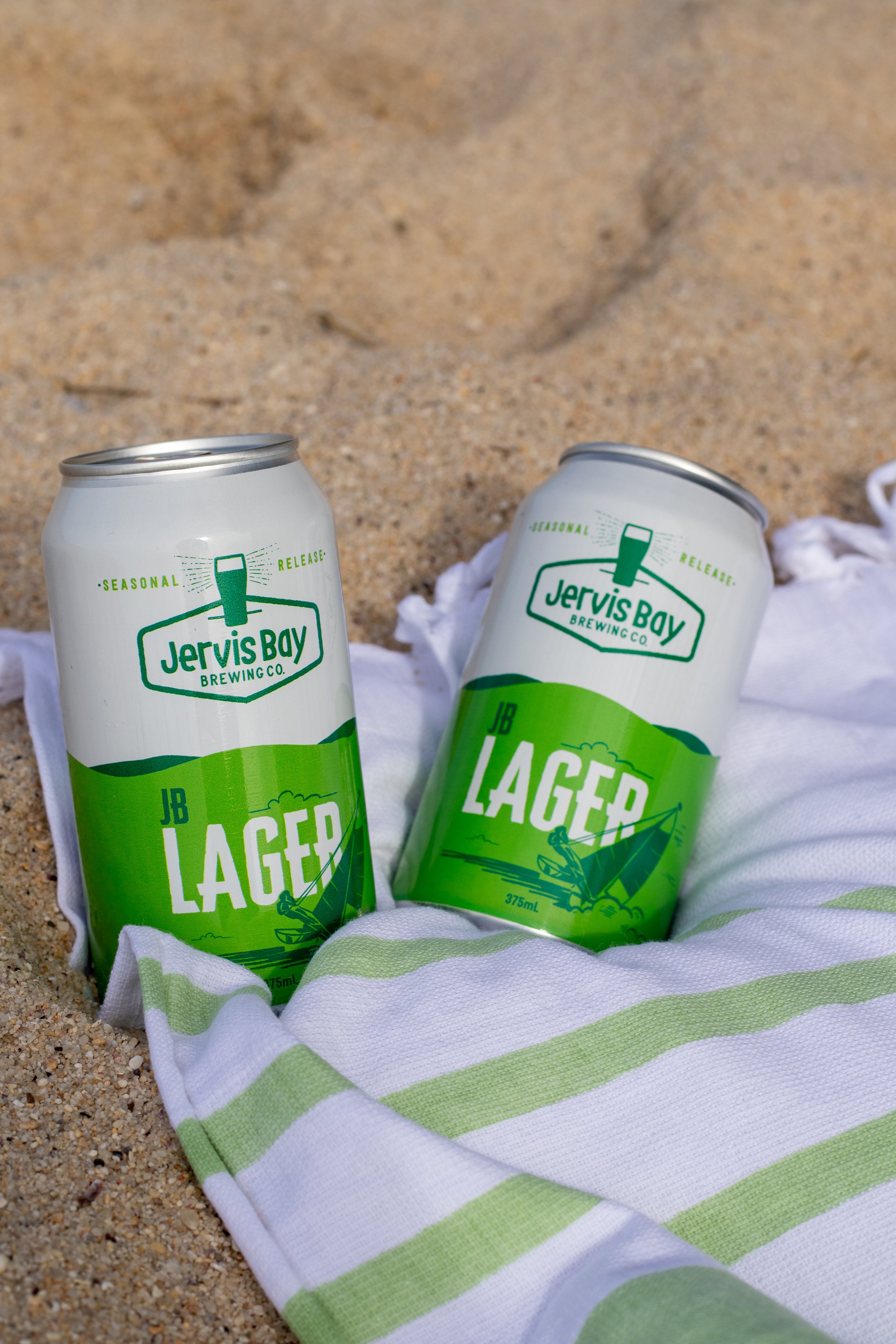 Two cans of Jervis Bay Brewing Co.'s JB Lager on sand, wrapped in a white towel with green stripes.