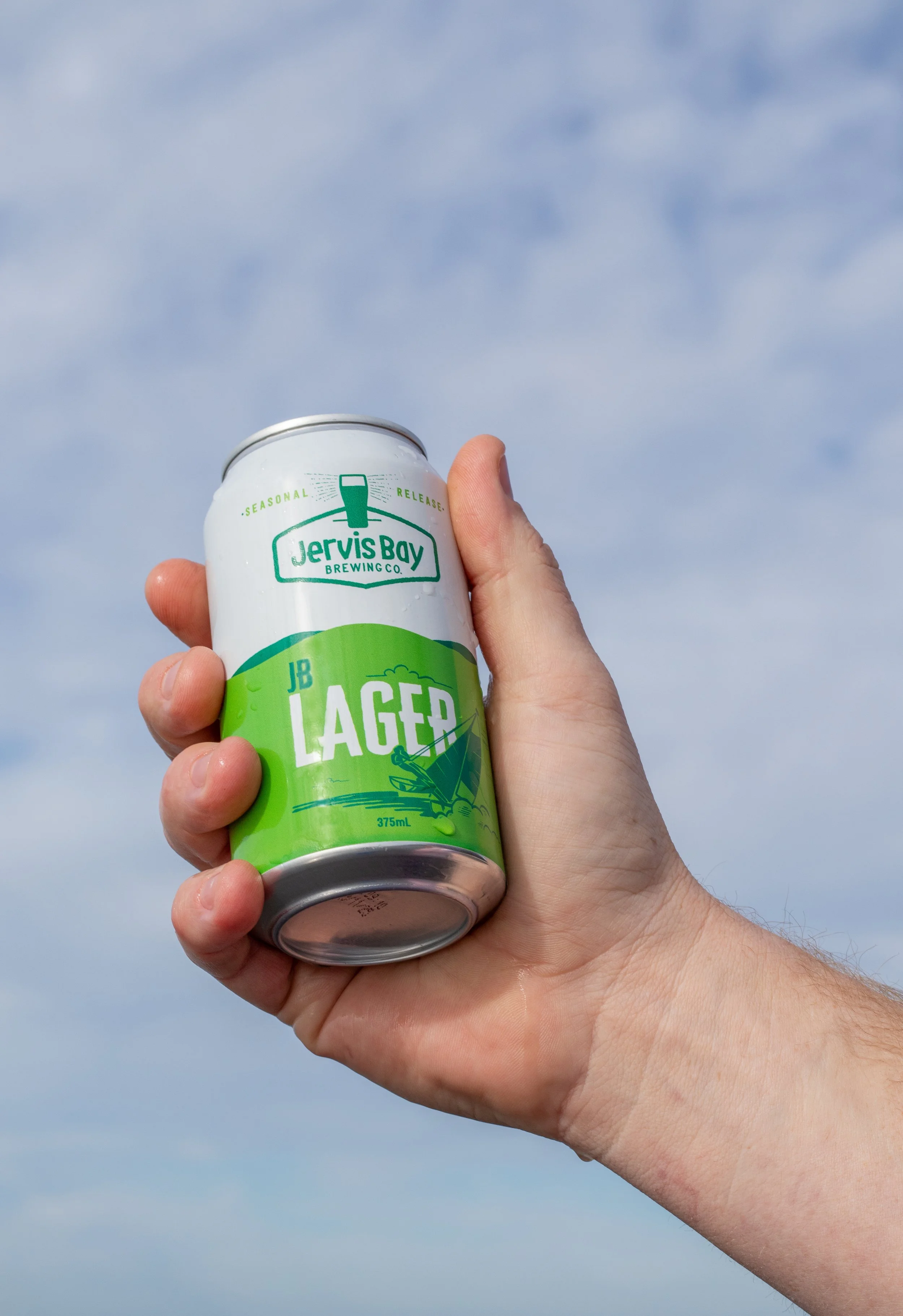 A hand holding a can of Jervis Bay Brewing JB Lager outdoors against a blue sky with clouds.