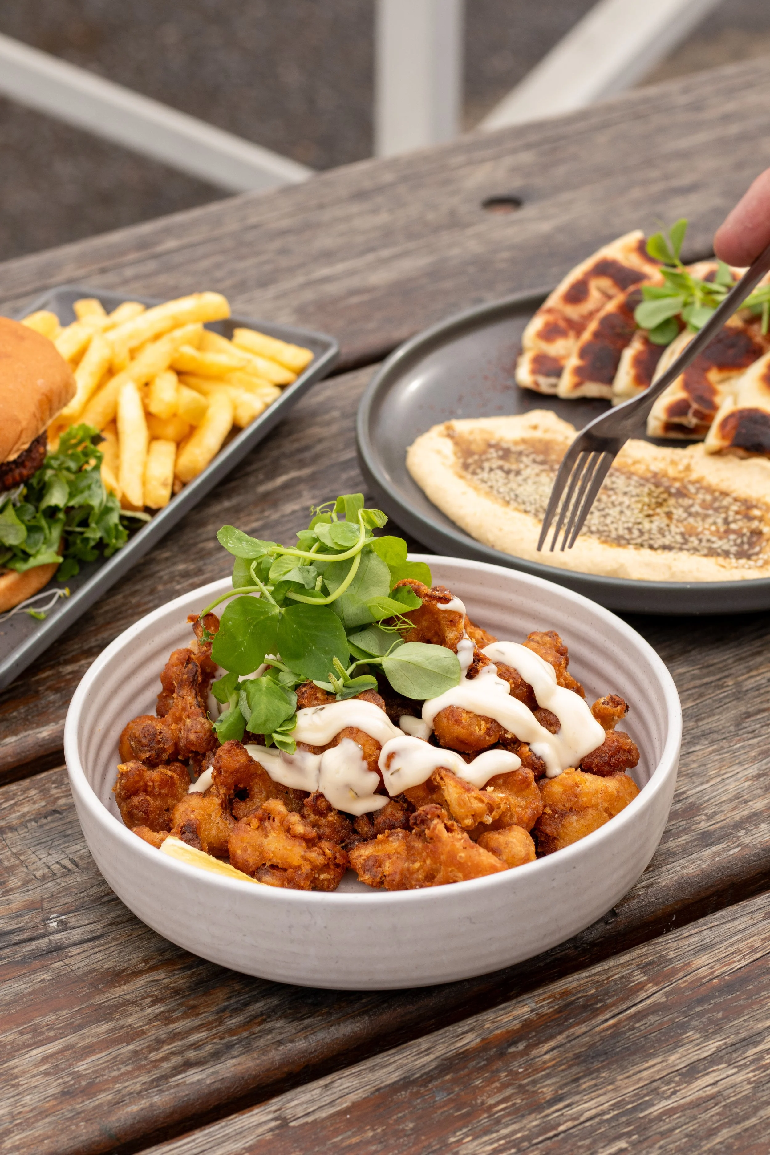 A bowl of crispy fried chicken topped with white sauce and garnished with fresh green herbs on a rustic wooden table, with a side of French fries and a plate of flatbread in the background.