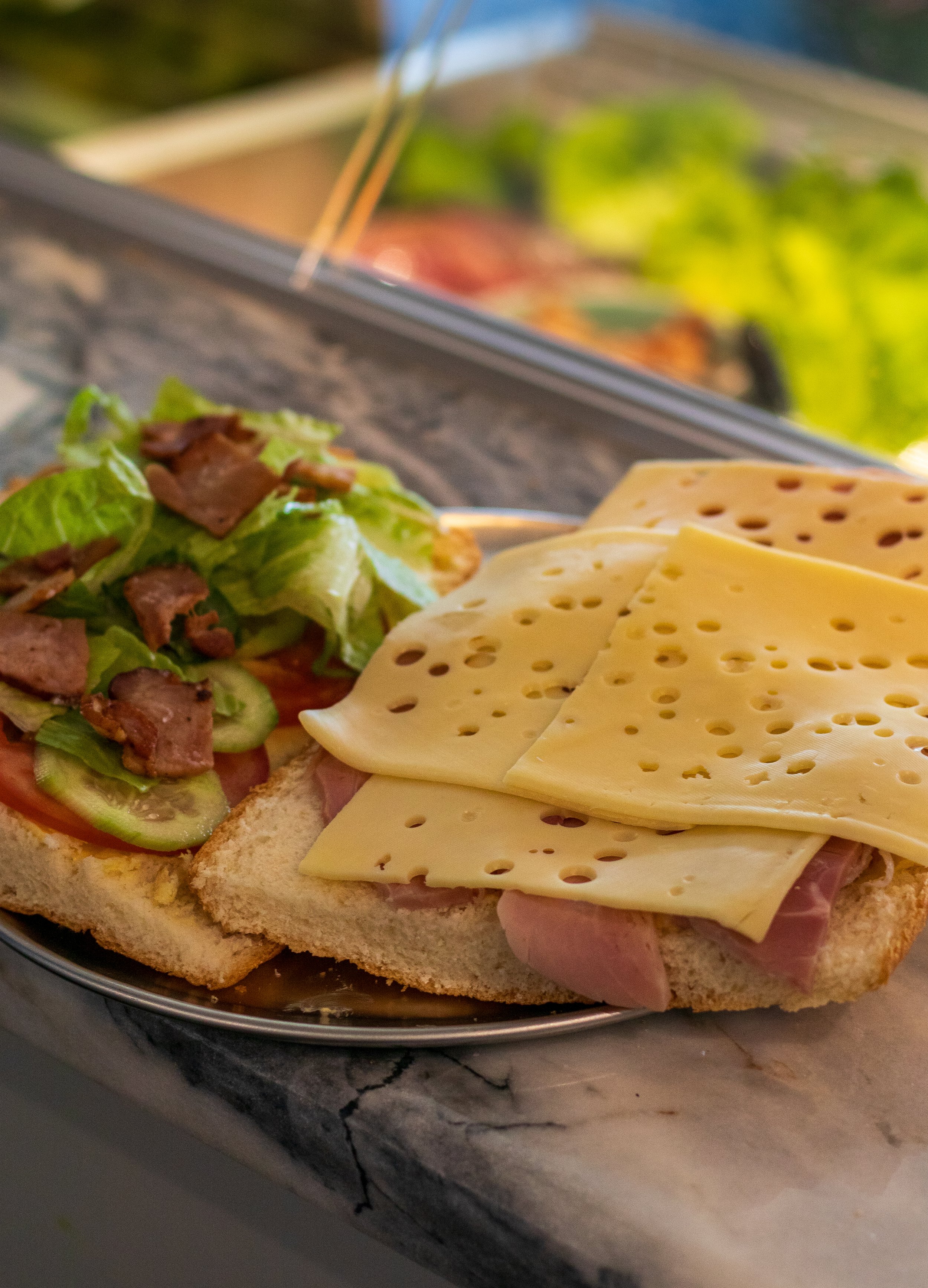 A sandwich with sliced cheese, ham, lettuce, tomato, cucumbers, and bacon on bread, served on a metal plate on a marble counter.