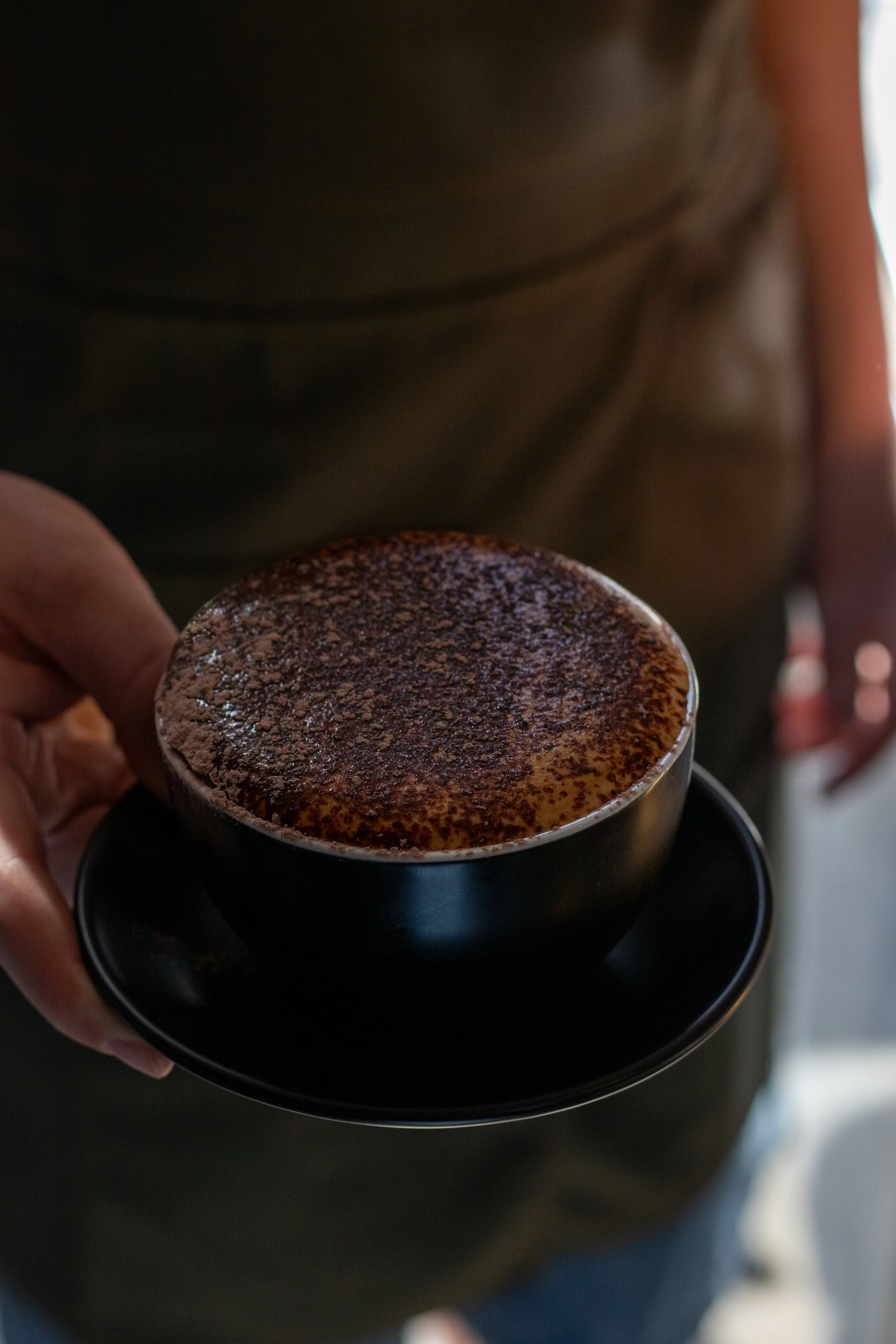 Person holding a black cup of cappuccino topped with cocoa powder on a black saucer.