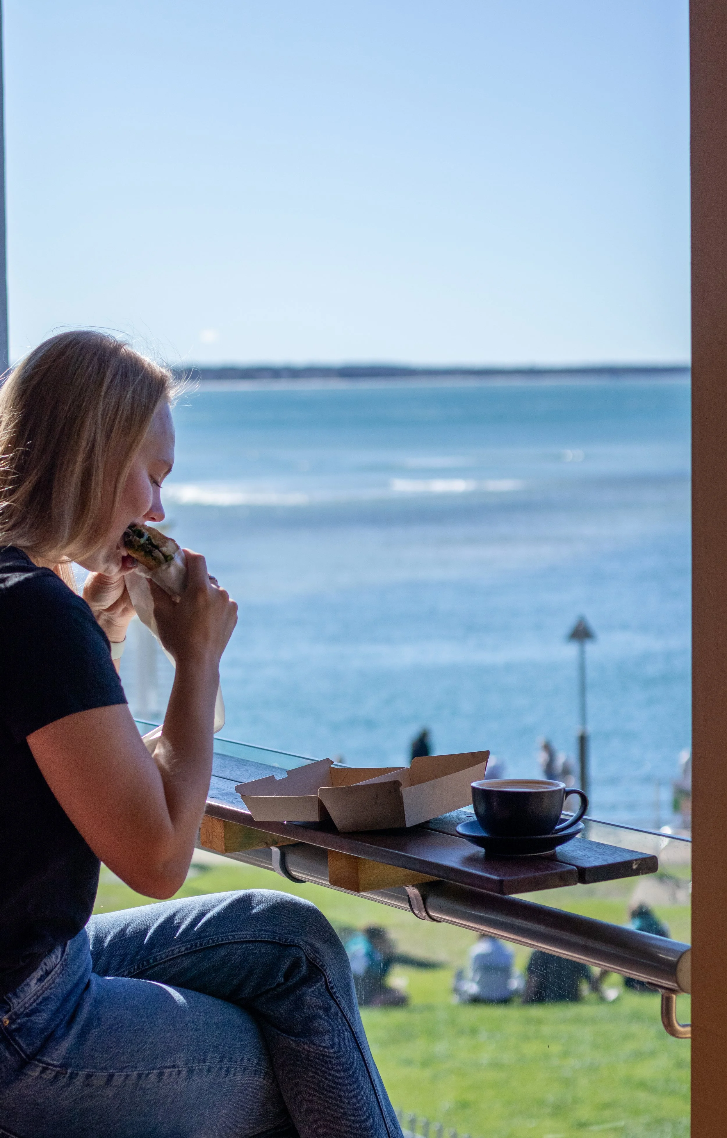 A woman sitting by a window with a view of the ocean, eating a sandwich, with a cup of coffee and a takeout box on the windowsill.