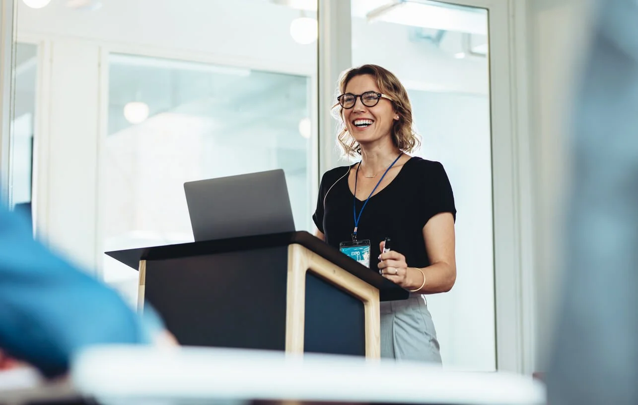A smiling woman with glasses giving a presentation at a podium in a modern office setting.