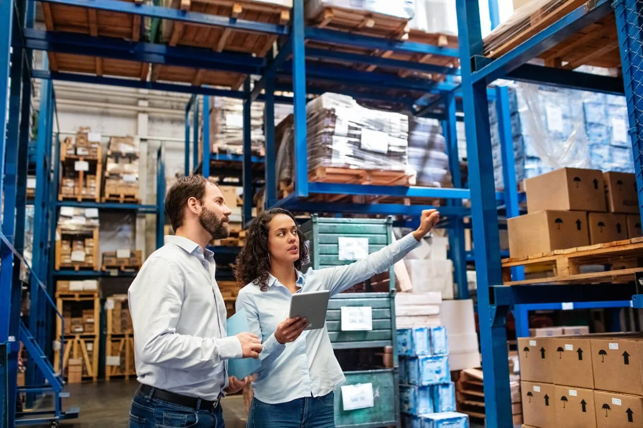 Two people, a man and a woman, are inspecting items on high shelves in a warehouse, with the woman pointing at a box while holding a tablet.