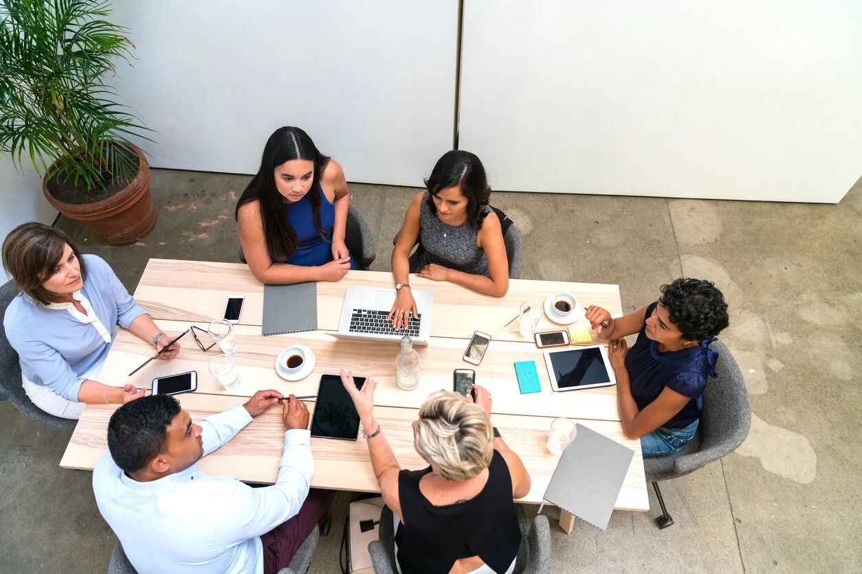 Six diverse women sitting around a rectangular table with laptops, tablets, and smartphones, engaged in a meeting or discussion.