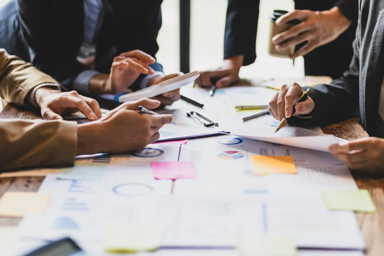 Group of people working on a business project at a table with documents, charts, and sticky notes.