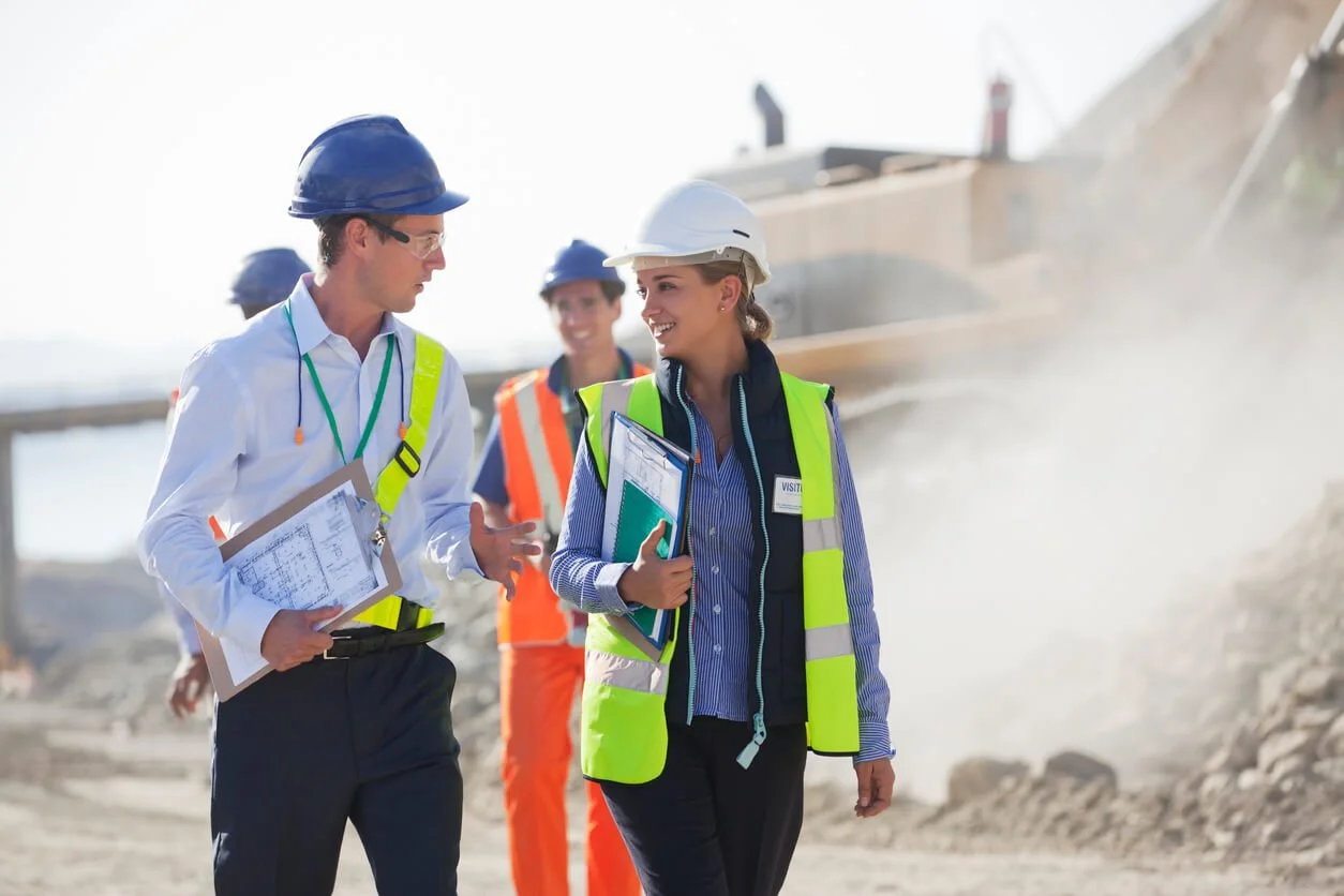 Construction workers wearing safety helmets and vests walking and talking at a construction site with machinery and dust in the background.
