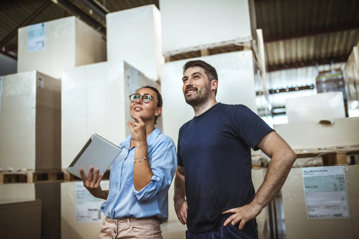 Two people, a woman with glasses holding a tablet and a man with a beard standing with hands on hips, in a warehouse with large boxes on shelves.
