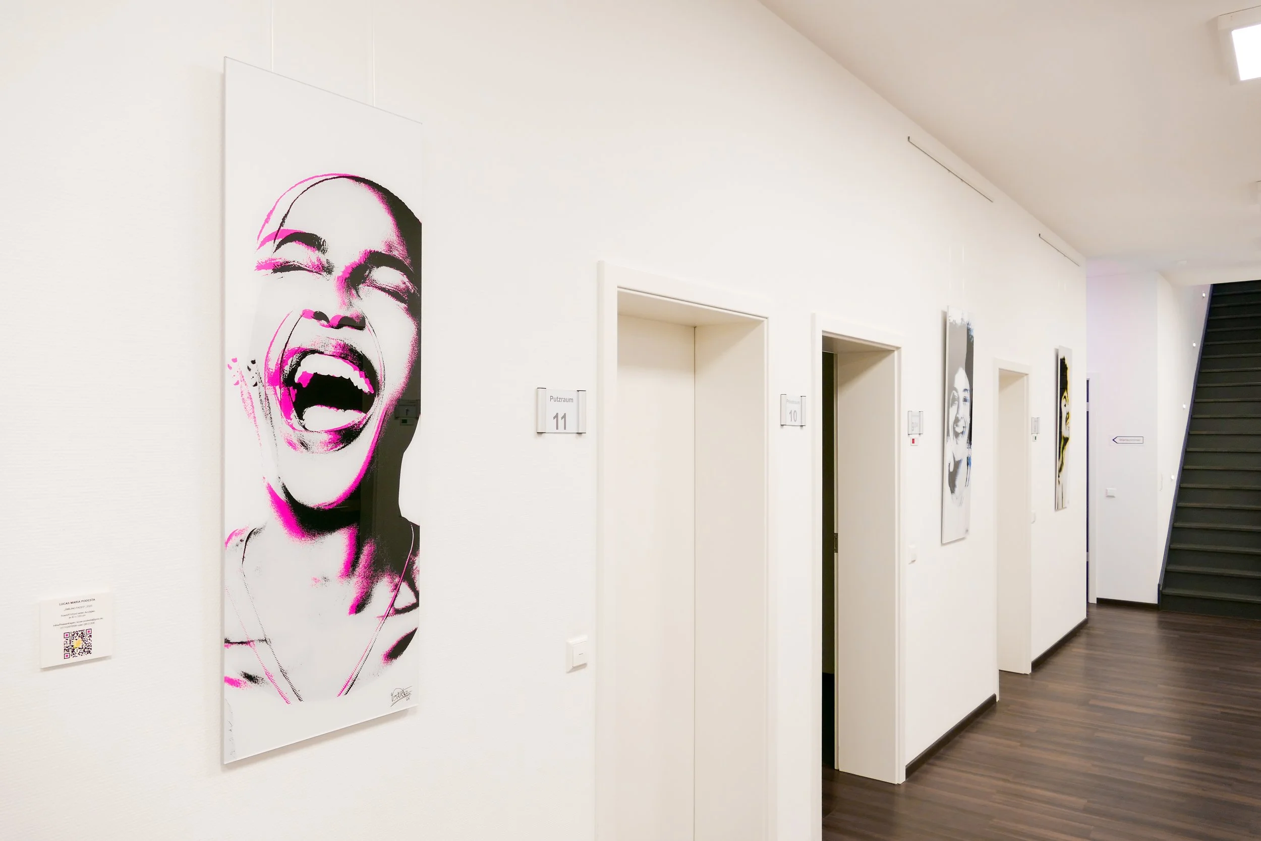 Weißer Flur mit mehreren Portraits von lachenden Frauen in Schwarz-Weiß mit pinken Akzentlinien, Türen mit Nummern 10 und 11, und eine Treppe im Hintergrund in einem modernen Gebäude.