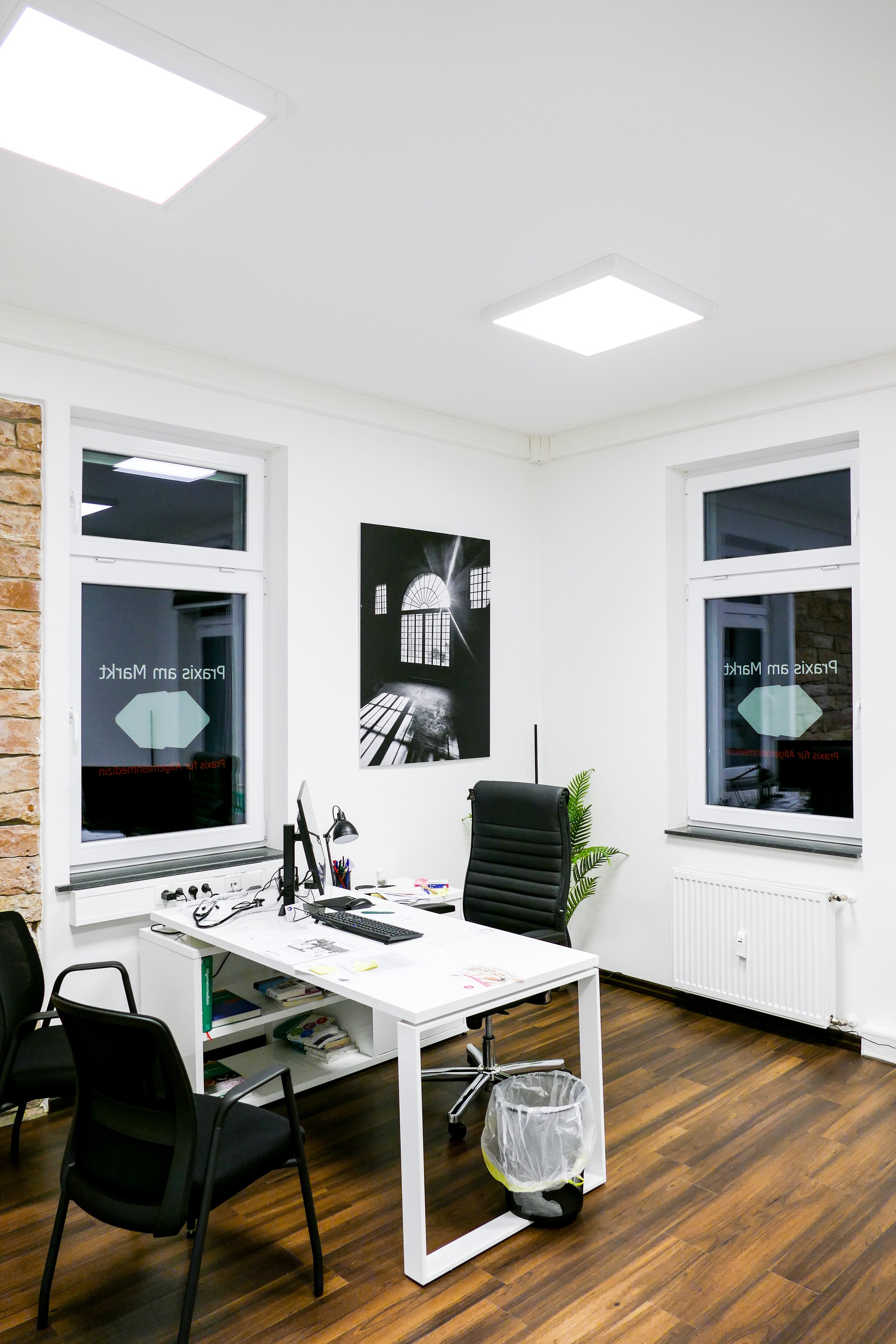 Ein modernes Büro mit weißem Tisch, schwarzem Bürostuhl, zwei schwarzen Besucherstühlen, einem Laptop, einem Bildschirm, Schreibutensilien, einer Pflanze, Wandkunst und großen Fenstern bei Nacht.