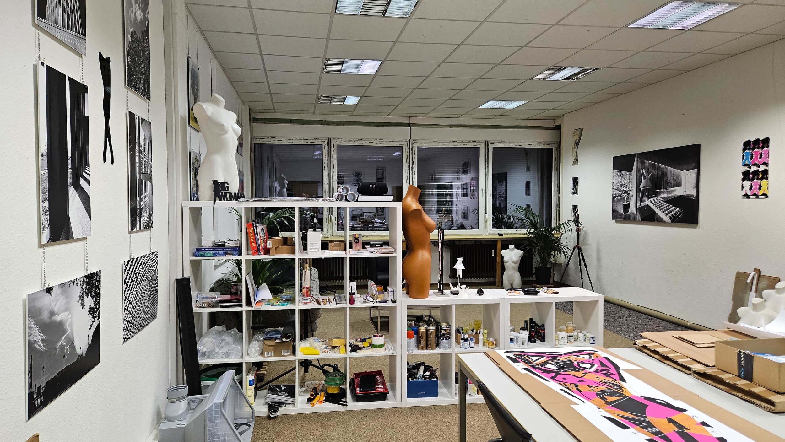 Kunststudio mit schwarz-weißen Fotos an den Wänden, Skulpturen, Malmaterialien und Werkzeugen auf Regalen und Tischen, große Fenster im Hintergrund, gedimmtes Kunstwerk an der Wand.