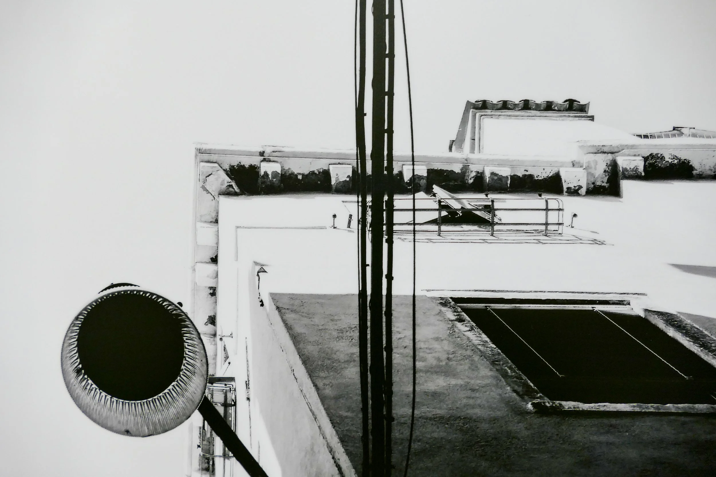 Schwarz-weiß Foto einer Hauswand mit einer Dachluke, Wasserleitung, Kabel, und einem runden Lüftungsschacht, aufgenommen von unten nach oben.