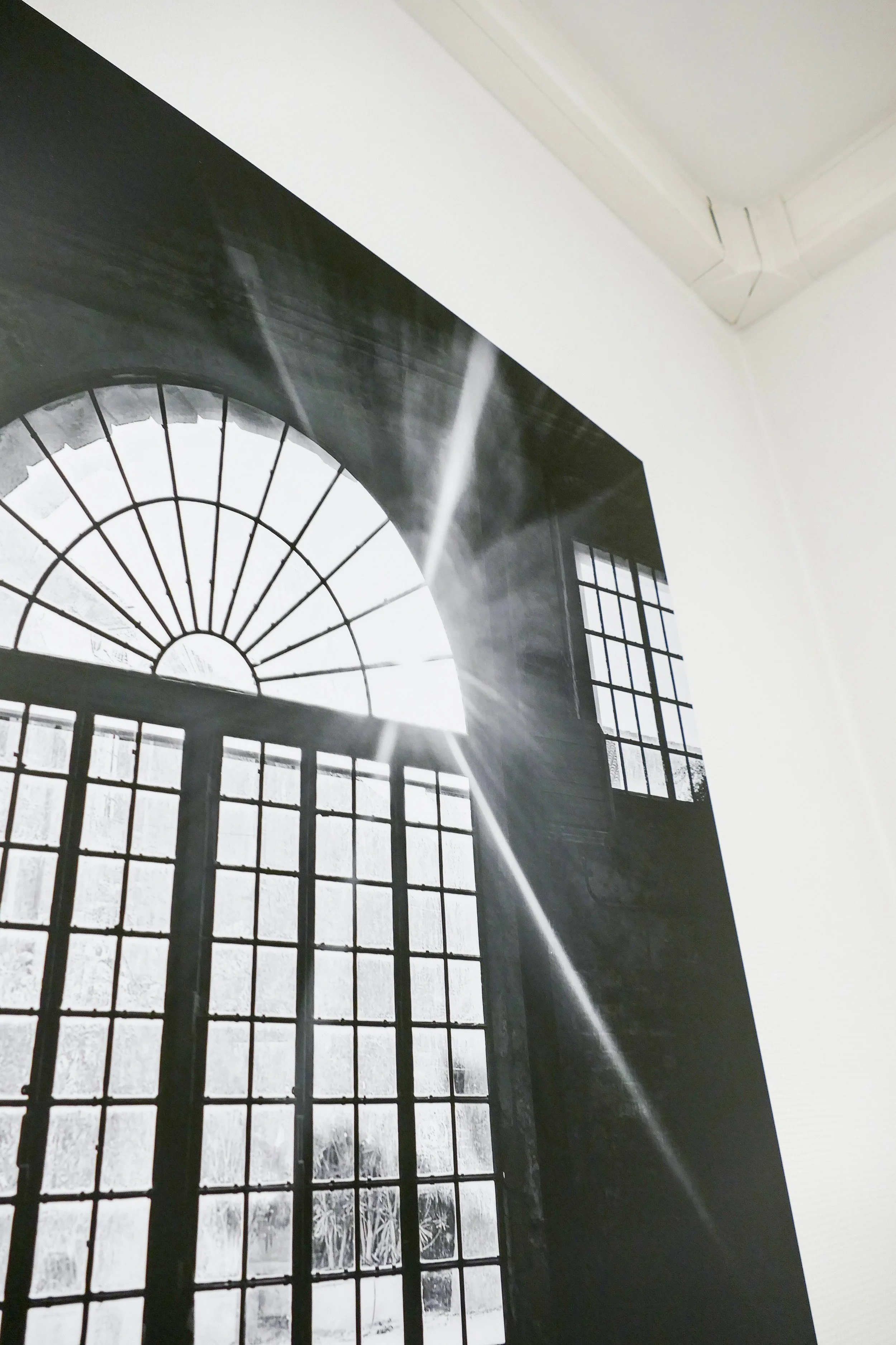 Schwarz-Weiß-Fotografie eines großen Fensters mit Bogen im Innenraum, Sonnenlicht scheint durch das Fenster und erzeugt Lichtstrahlen.