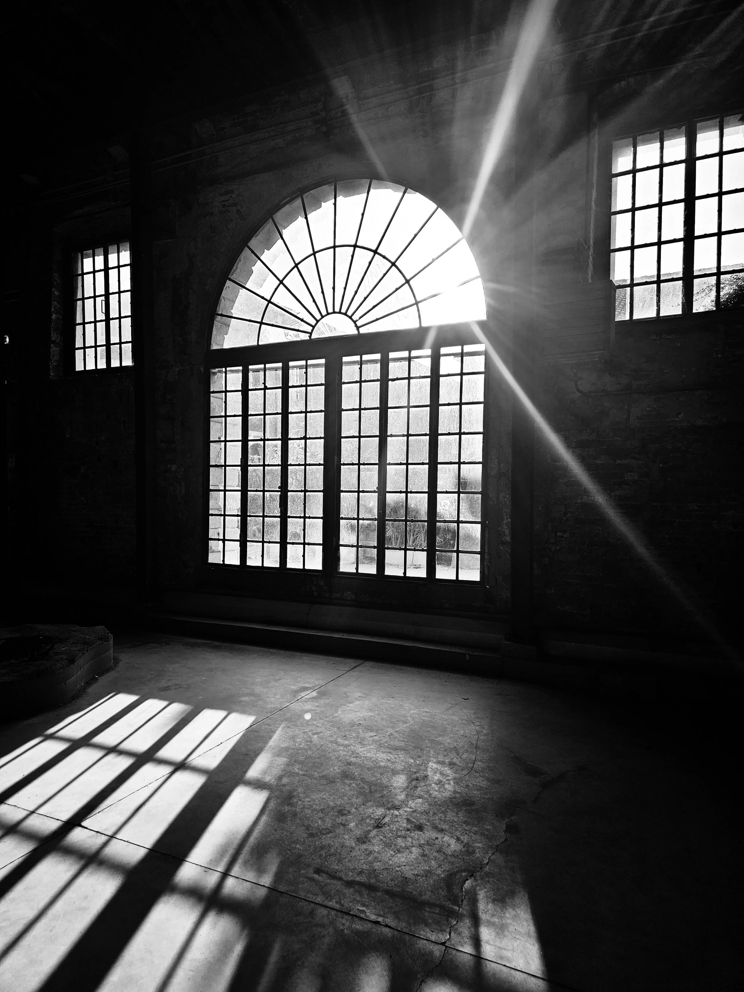 Schwarz-weiß-Foto eines alten Gebäudes mit großen Fenstern, Sonnenstrahlen, die durch die Fenster scheinen, und Schatten auf dem Boden.