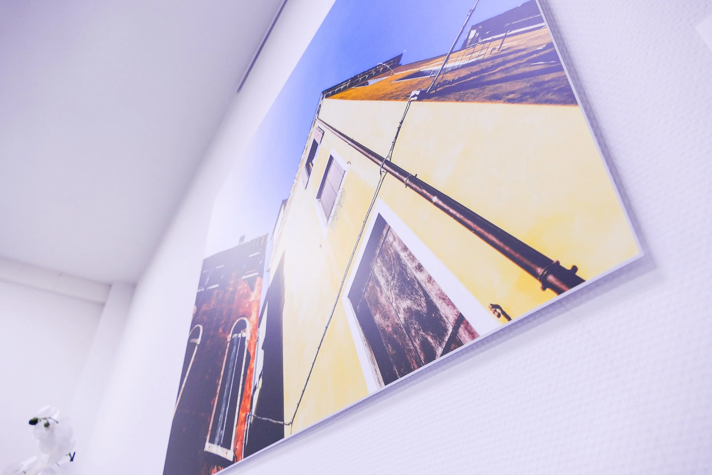 Fotografie eines Gebäudes mit gelber Wand, Fenster und schwarzen Rohren, an einer weißen Wand in einem Raum aufgehängt