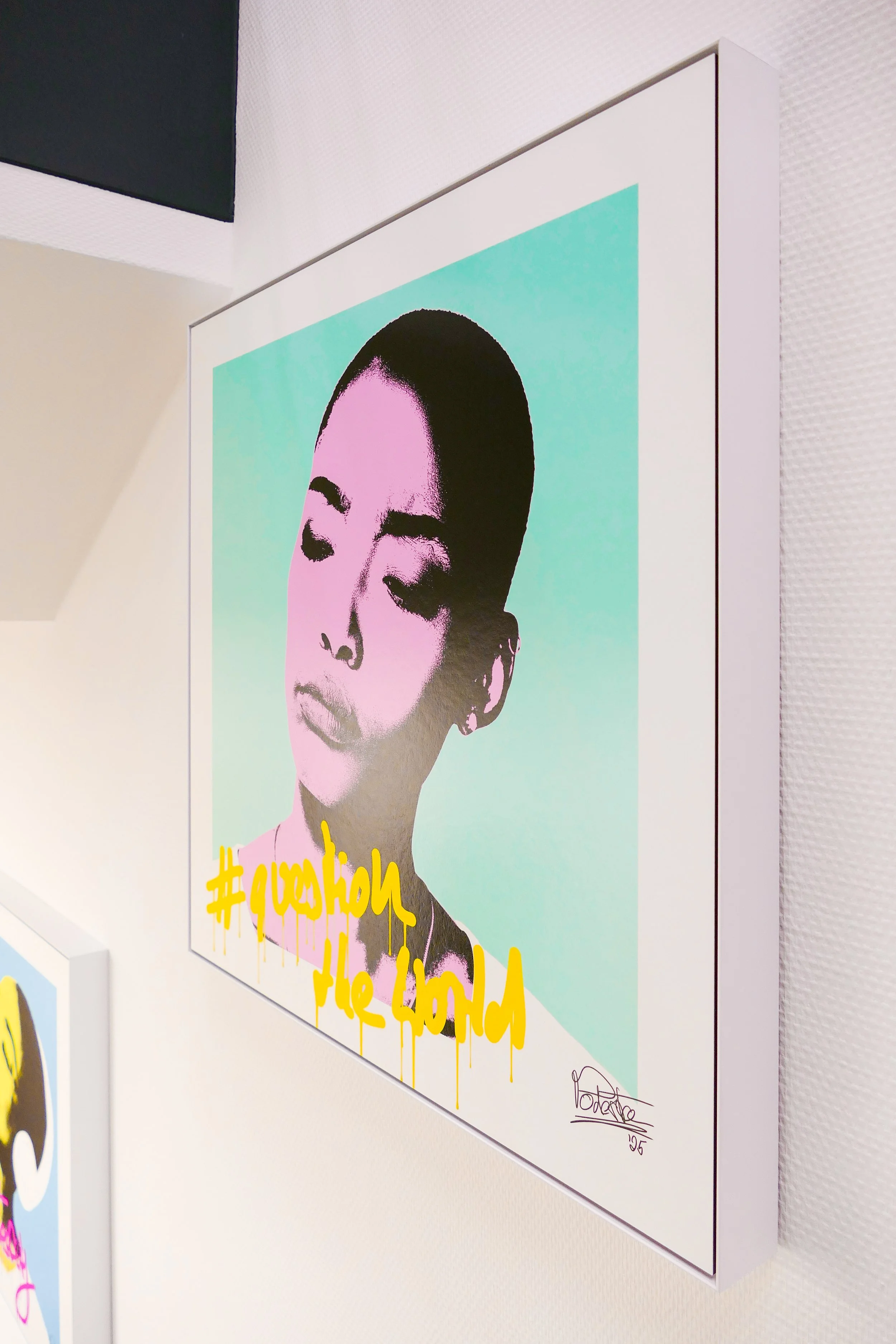 Ein modernes Pop-Art-Porträt einer Frau mit geschlossenen Augen. Darunter befindet sich gelbe Graffiti-Schrift, die sagt '#question the world'. Das Bild ist in einem weißen Rahmen und hängt an einer weißen Wand, die einen schwarzen Abschnitt hat.