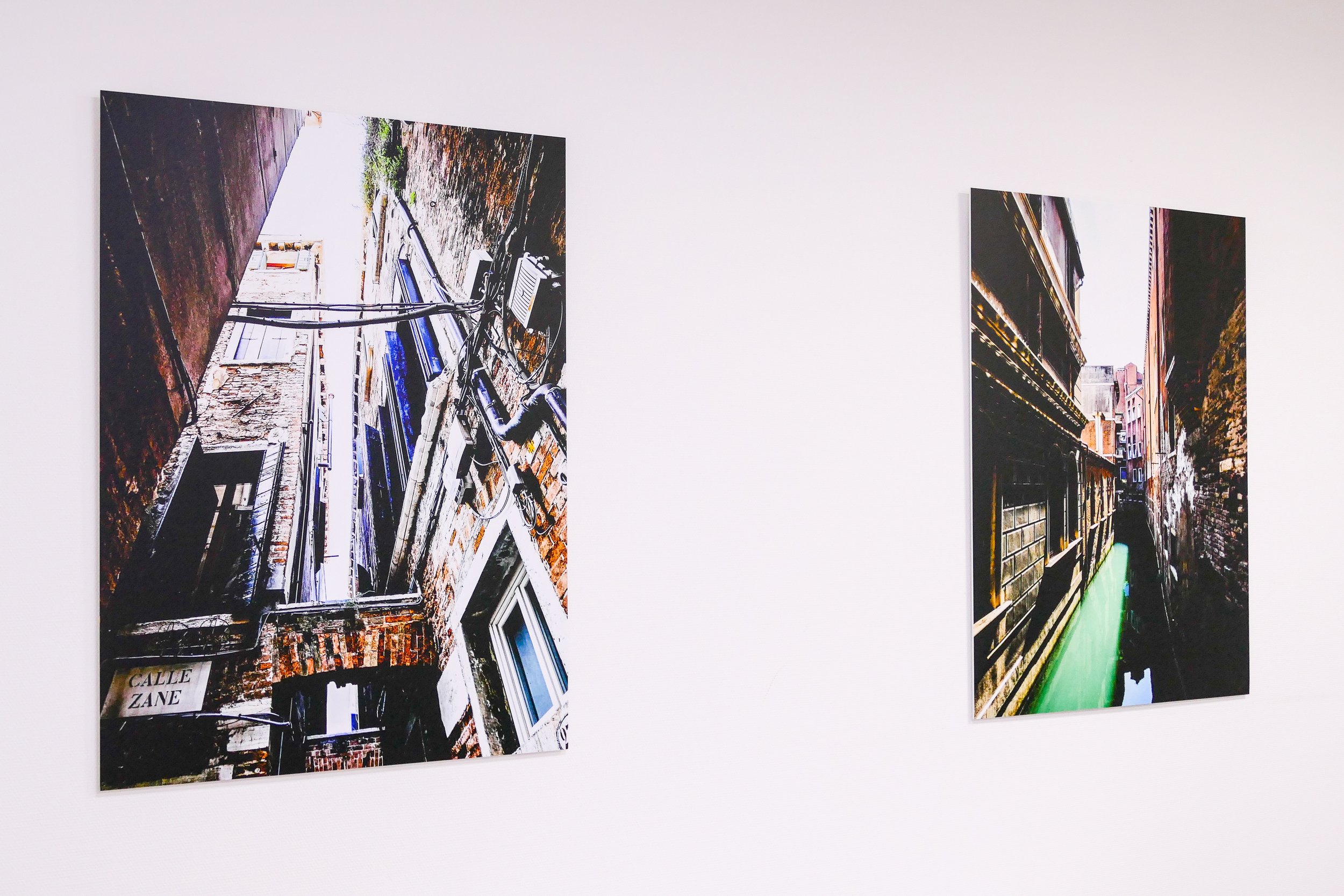 Zwei Fotografien, die an einer weißen Wand hängen, zeigen enge Kanäle in einer Stadt. Das Bild auf der linken Seite zeigt eine Gebäudeansicht mit Kabeln, während das rechte Bild einen Kanal mit Gebäuden auf beiden Seiten zeigt.