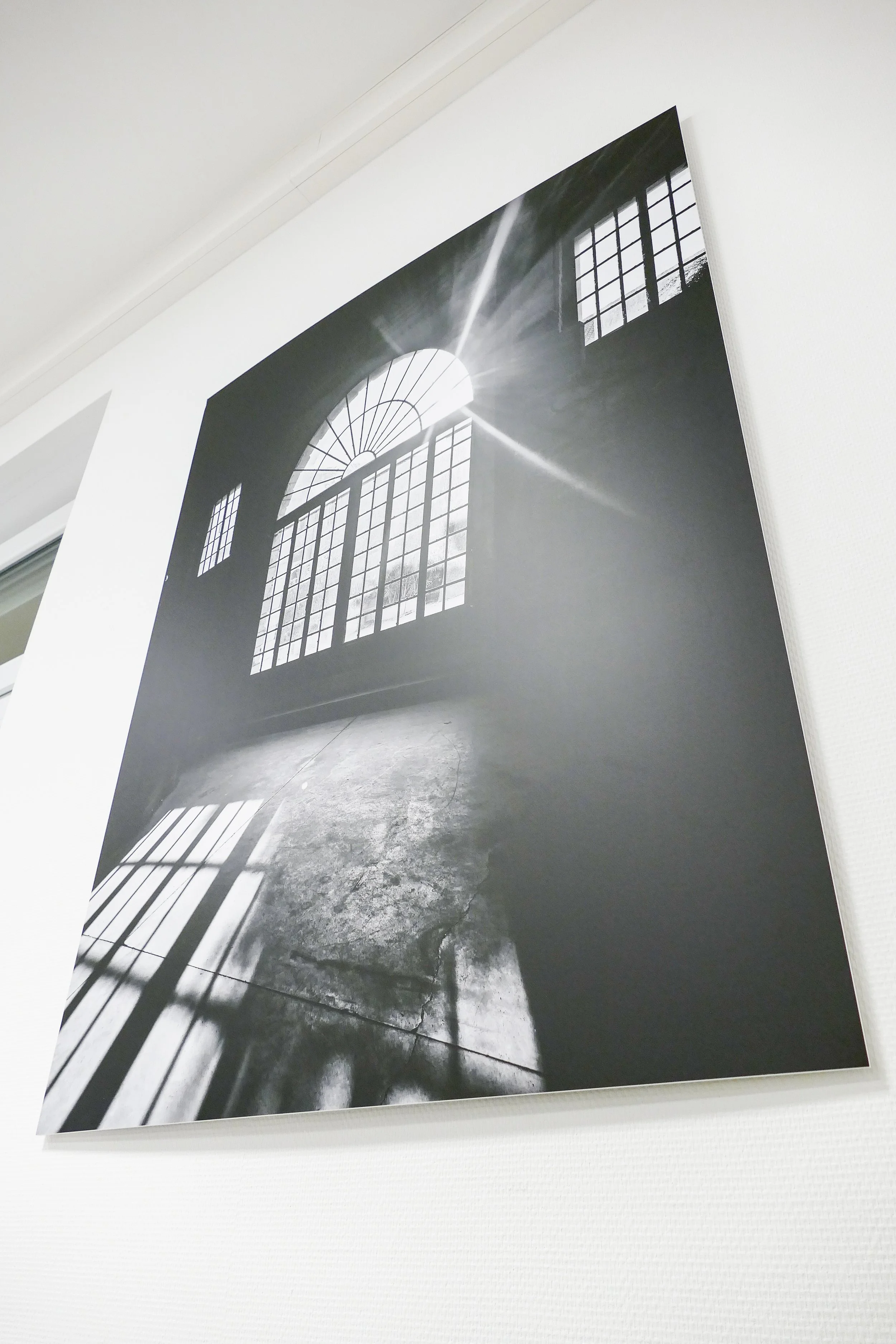 Schwarz-weiß-Fotografie eines großen Fensters mit Sonnenlicht, das in einen leeren Raum mit staubigem Boden scheint.