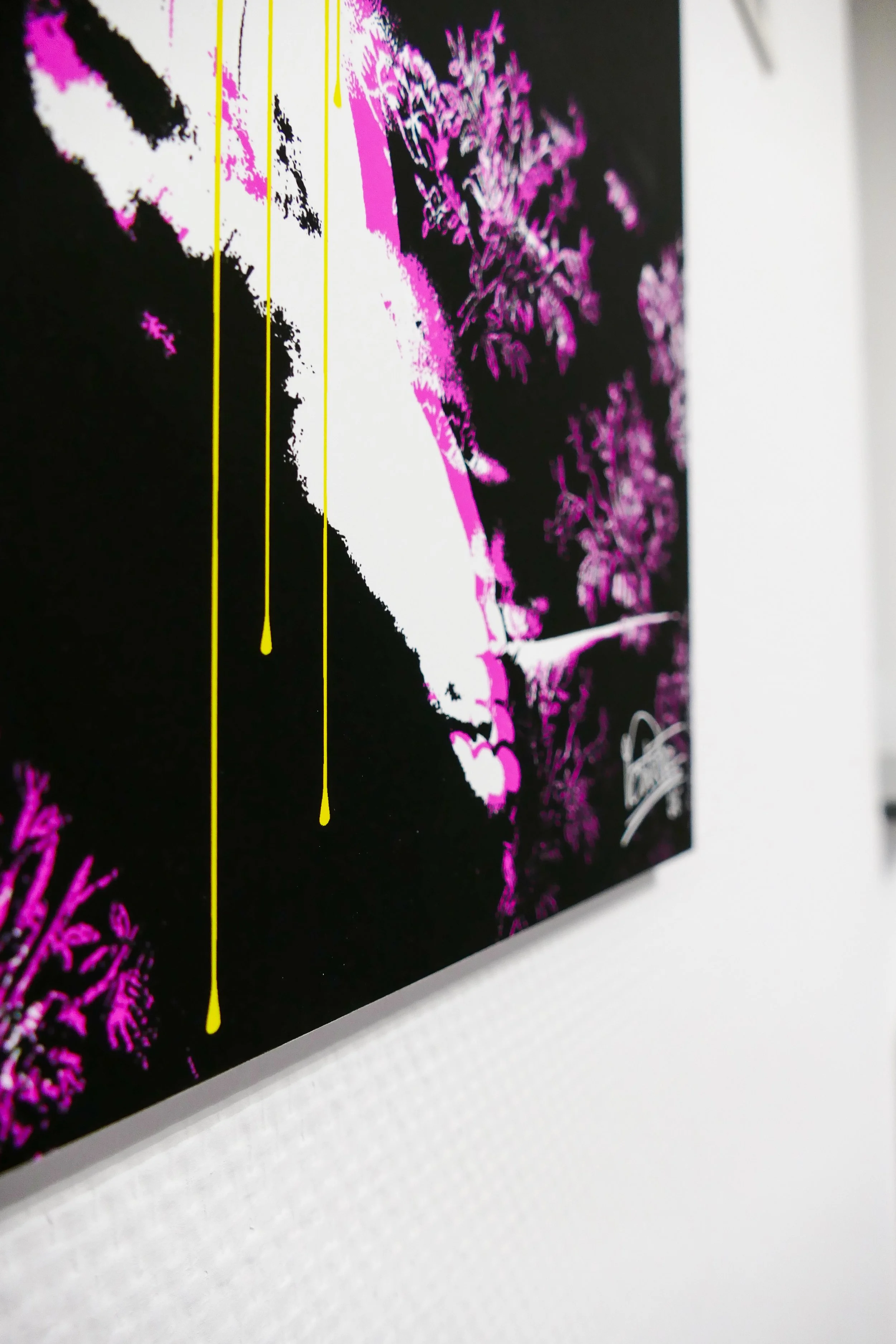 Ein Kunstwerk auf einer schwarzen Leinwand mit pinken und weißen Akzenten, darunter gelbe Tropfen, an einer weißen Wand.