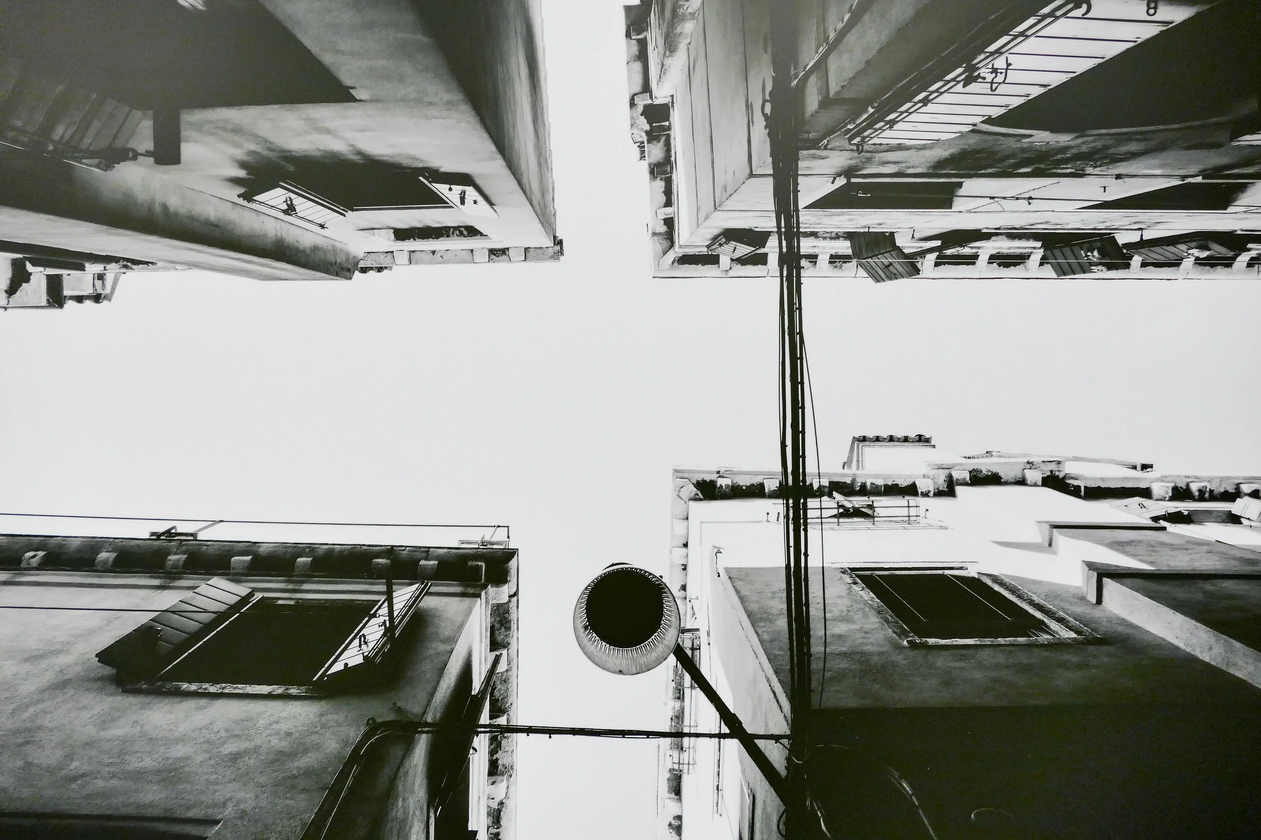 Schwarz-weiß-Foto, Blick nach oben zwischen mehreren alten, mehrstöckigen Gebäuden mit Fenstern und Türen, mit einem runden Lampenschirm und Kabeln in der Mitte.