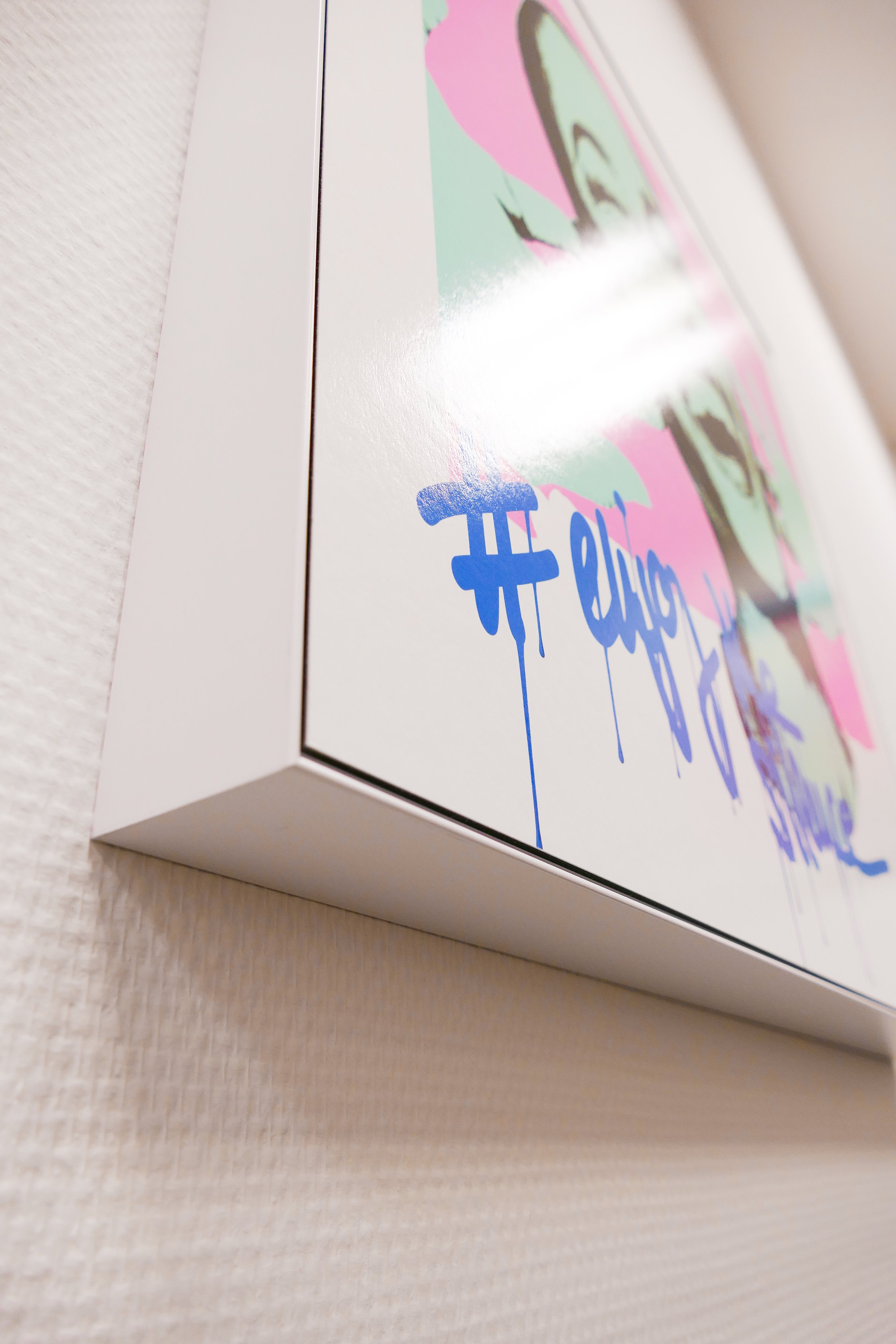 Das Bild zeigt eine Nahaufnahme eines weißen Rahmens mit einer Illustration, die ein buntes Bild mit pink, grün und schwarzen Elementen enthält, sowie blauen graffitiartigen Text.