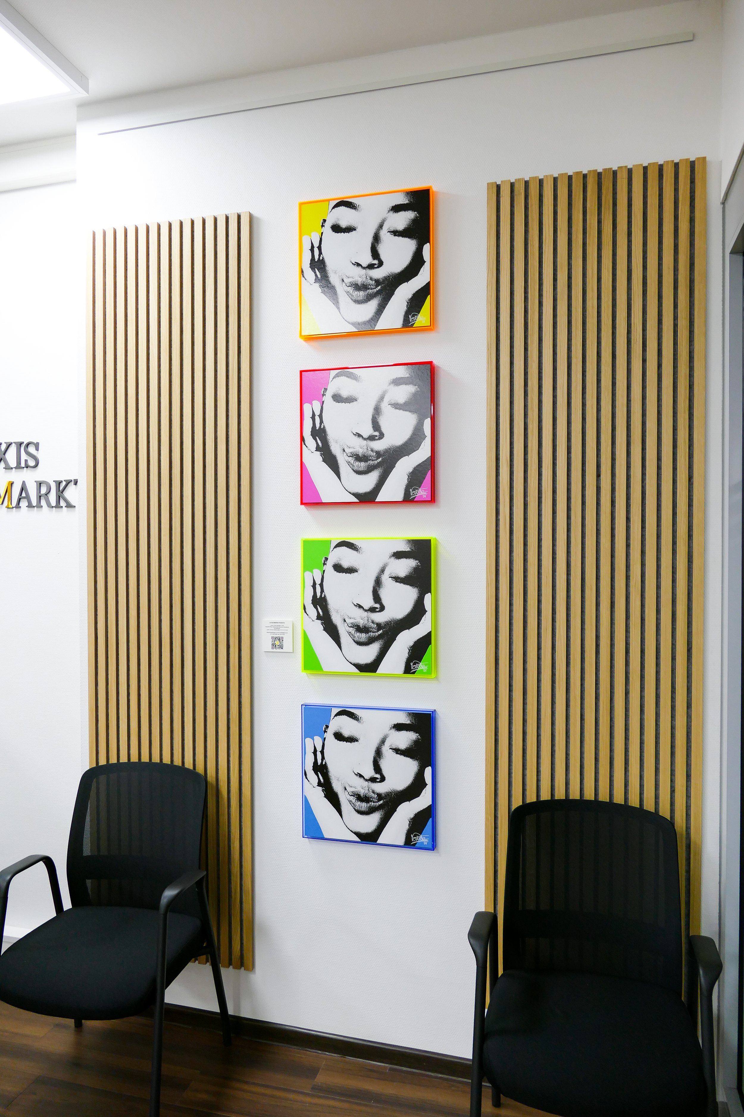 Vier Pop-Art-Bilder eines lachenden Gesichts, in verschiedenen Farben umrahmt, hängen an einer weißen Wand, flankiert von Holzlamellen. Zwei schwarze Stühle stehen vor der Wand.