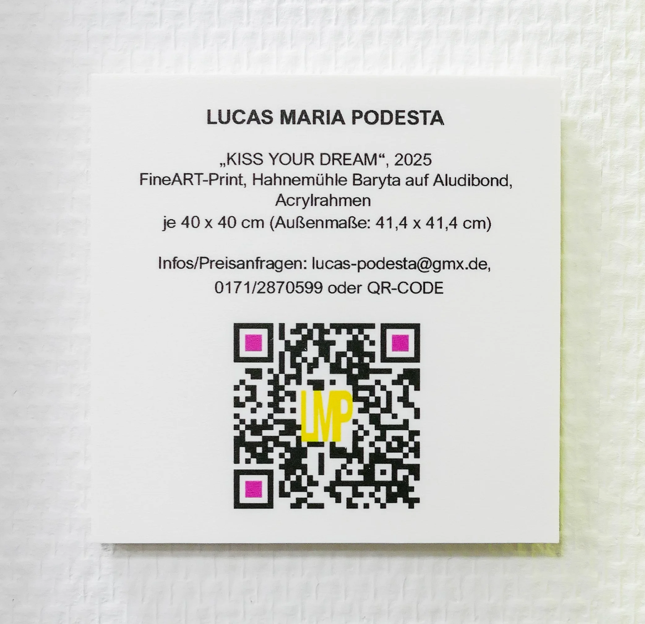 Ein weißes Blatt mit schwarzem QR-Code, pinken Ecken und gelbem Buchstaben 'IMP' in der Mitte. Oben steht der Name 'LUCAS MARIA PODESTA' und darunter Informationen zu einem Kunstwerk namens 'KISS YOUR DREAM', 2025, sowie Kontaktinformationen.