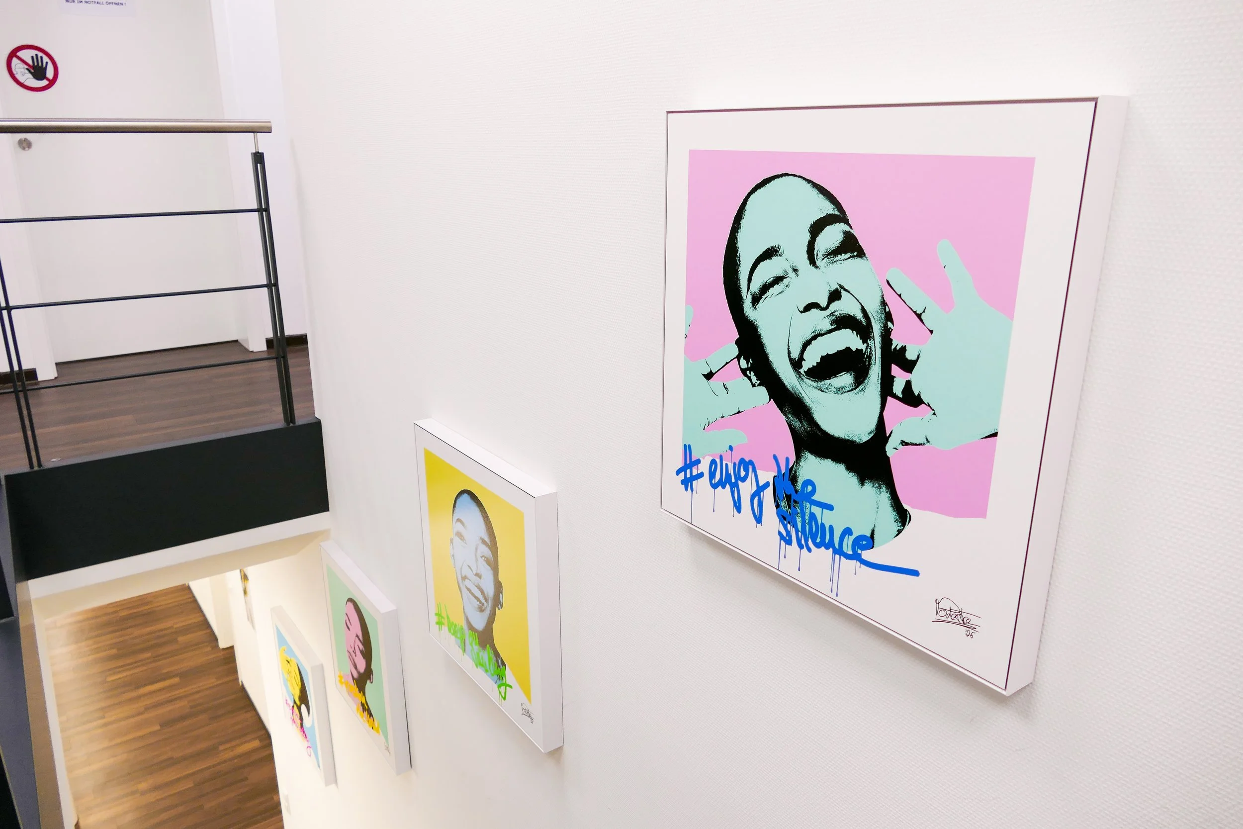 Mehrere kunstvolle Porträts in Pop-Art-Stil an einer weißen Wand. Das nächstgelegene Bild zeigt eine lachende Frau mit offenem Mund und Händen, die sie sich an die Ohren hält, mit pinkem Hintergrund und blauer Graffiti-Schrift darunter.