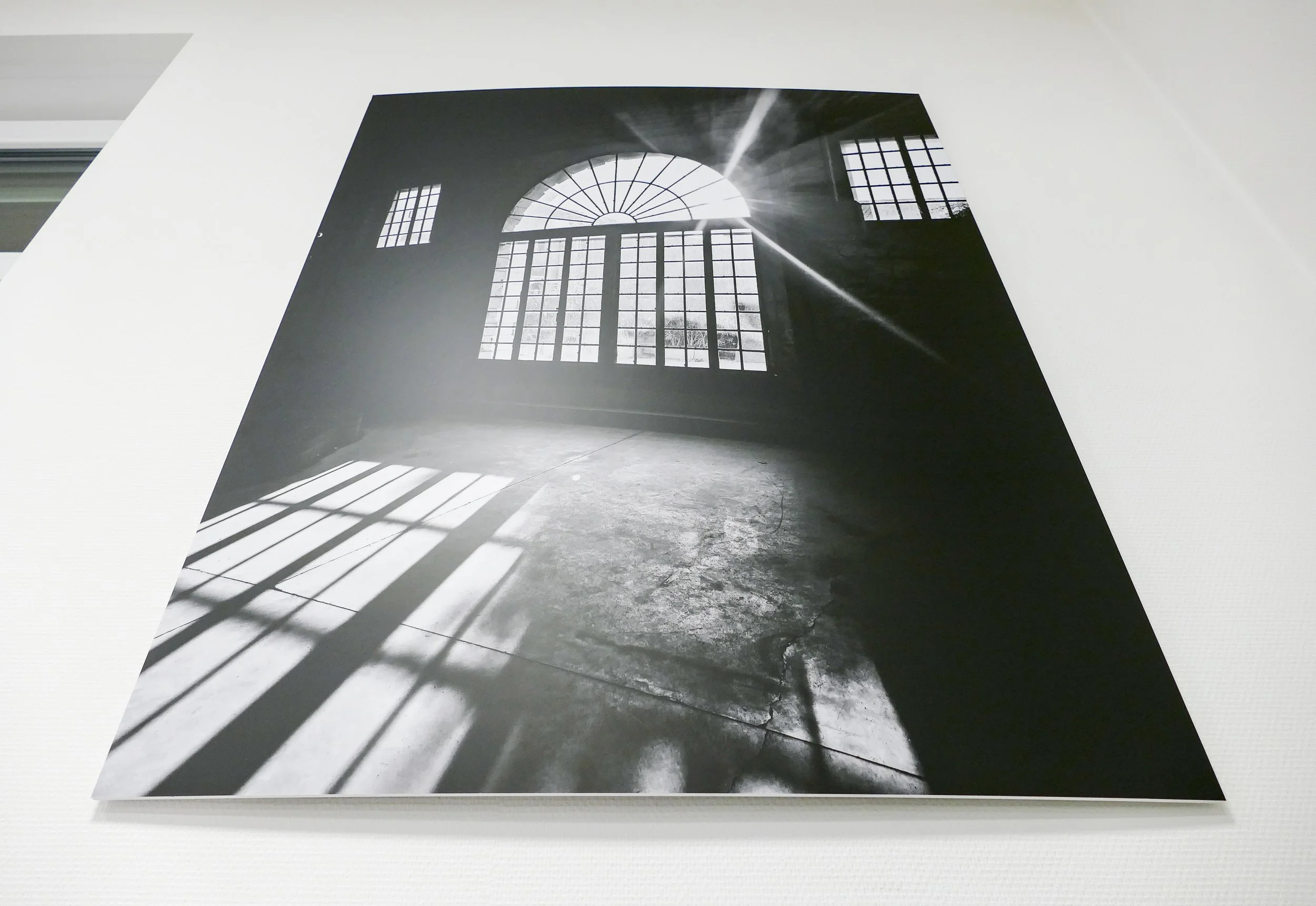 Schwarze und weiße Fotografie eines Raumes mit großen Fenstern, durch die Sonnenlicht und Schatten auf den Boden fallen.