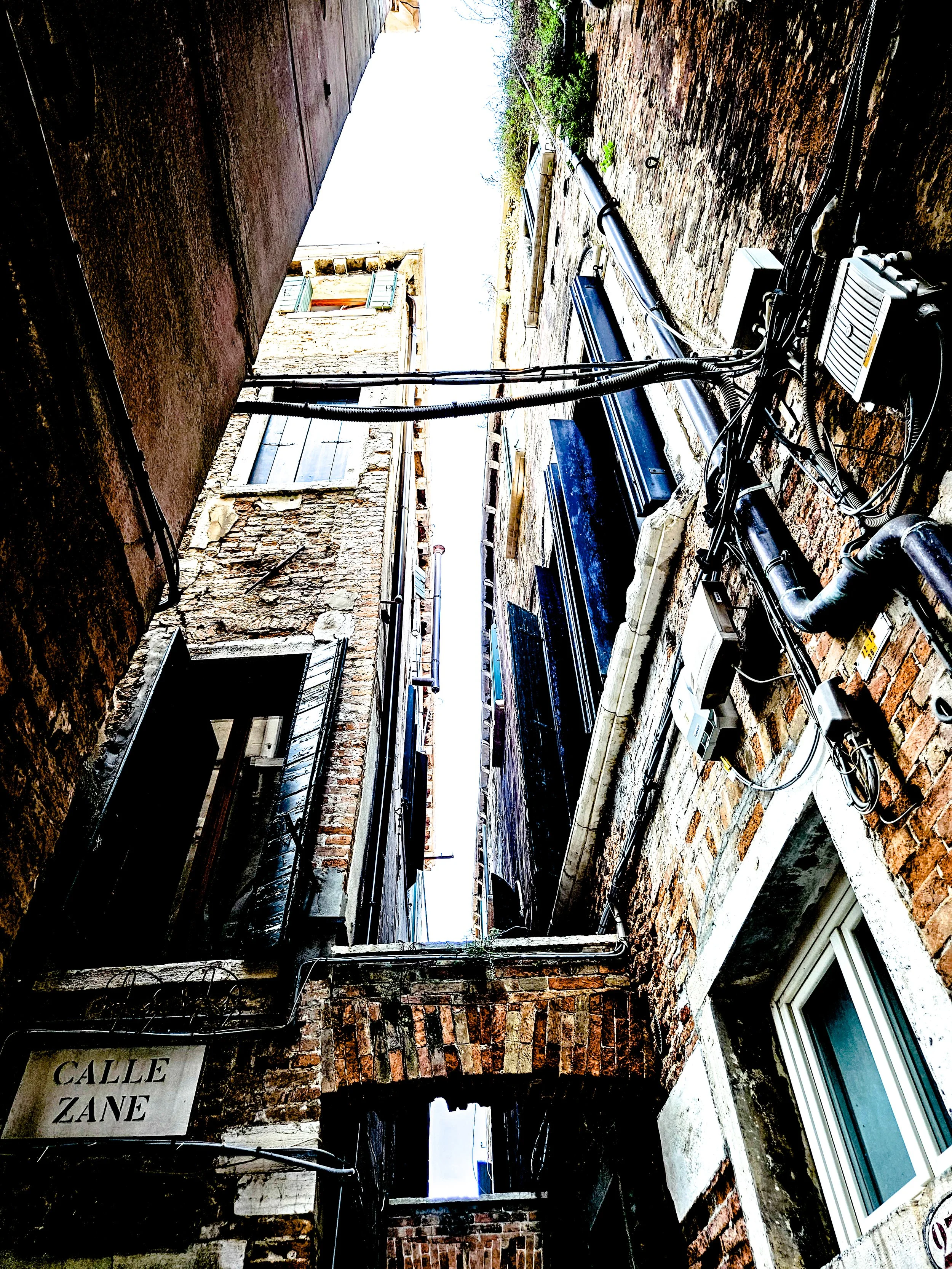 Blick nach oben in eine enge Gasse mit alten Backsteinbauten, Stromleitungen und Fenster.