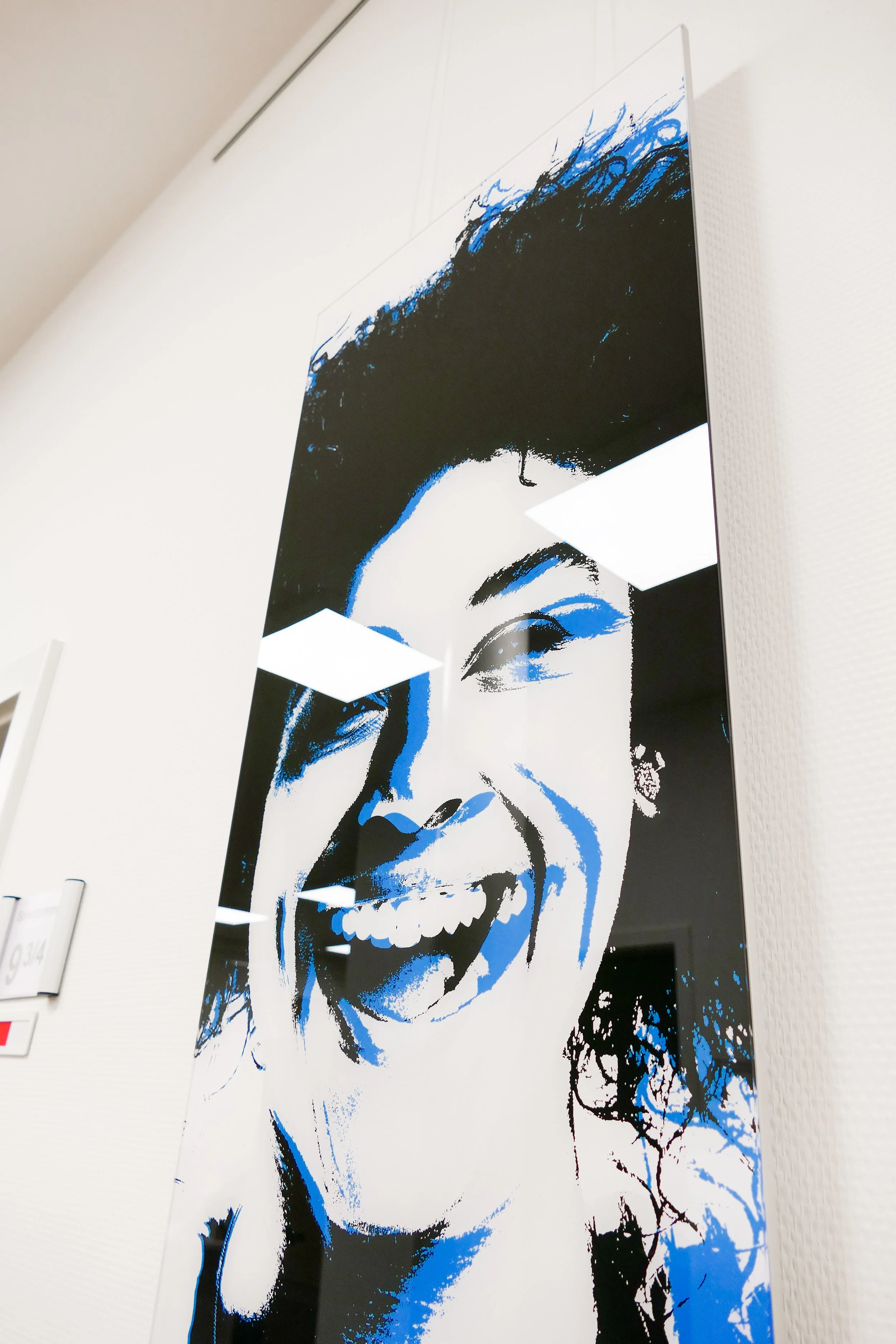 Ein großes, modernes Kunstwerk eines lachenden Gesichts, bestehend aus schwarzen, blauen und weißen Farben, hängt an einer weißen Wand in einem Innenraum.