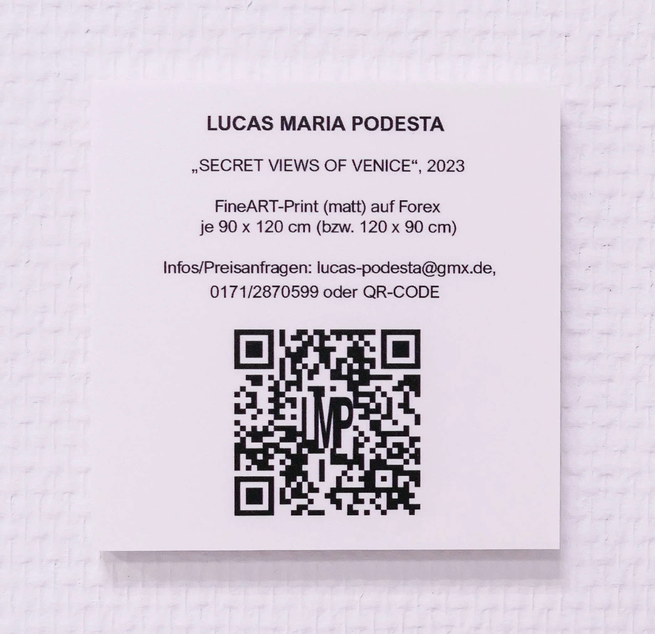 Ein weißer Zettel mit schwarzem Text und QR-Code. Text enthält Informationen über eine Kunst-Ausstellung von Lucas Maria Podesta, Titel 'Secret Views of Venice', 2023, mit Bildgröße 90x120 cm oder 120x90 cm, Kontaktinformationen und QR-Code.
