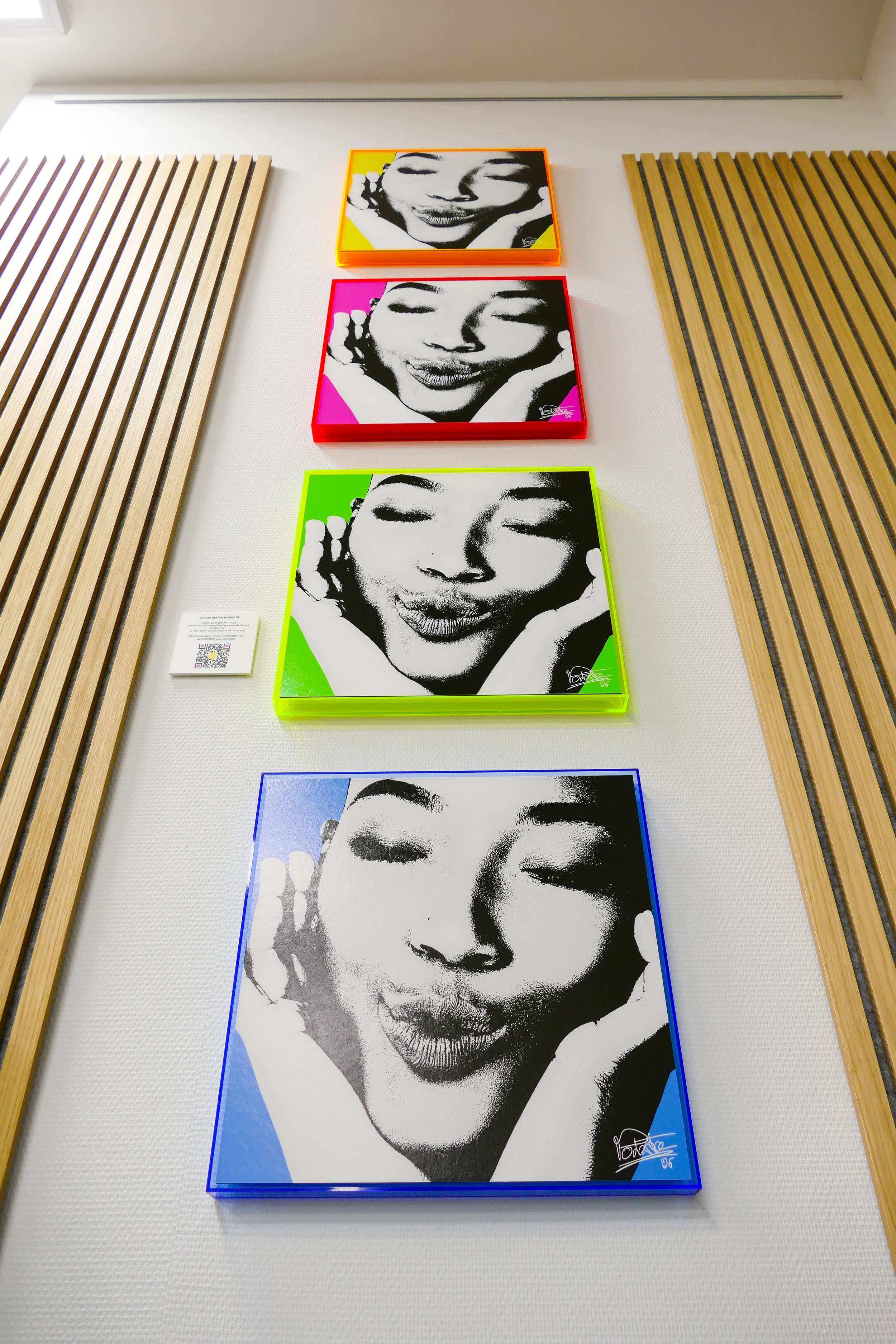 Vier farbige Kunstwerke einer lachenden Frau mit geschlossenen Augen, präsentiert in Frames mit verschiedenen Farben, an einer weißen Wand neben Holzpanelen.
