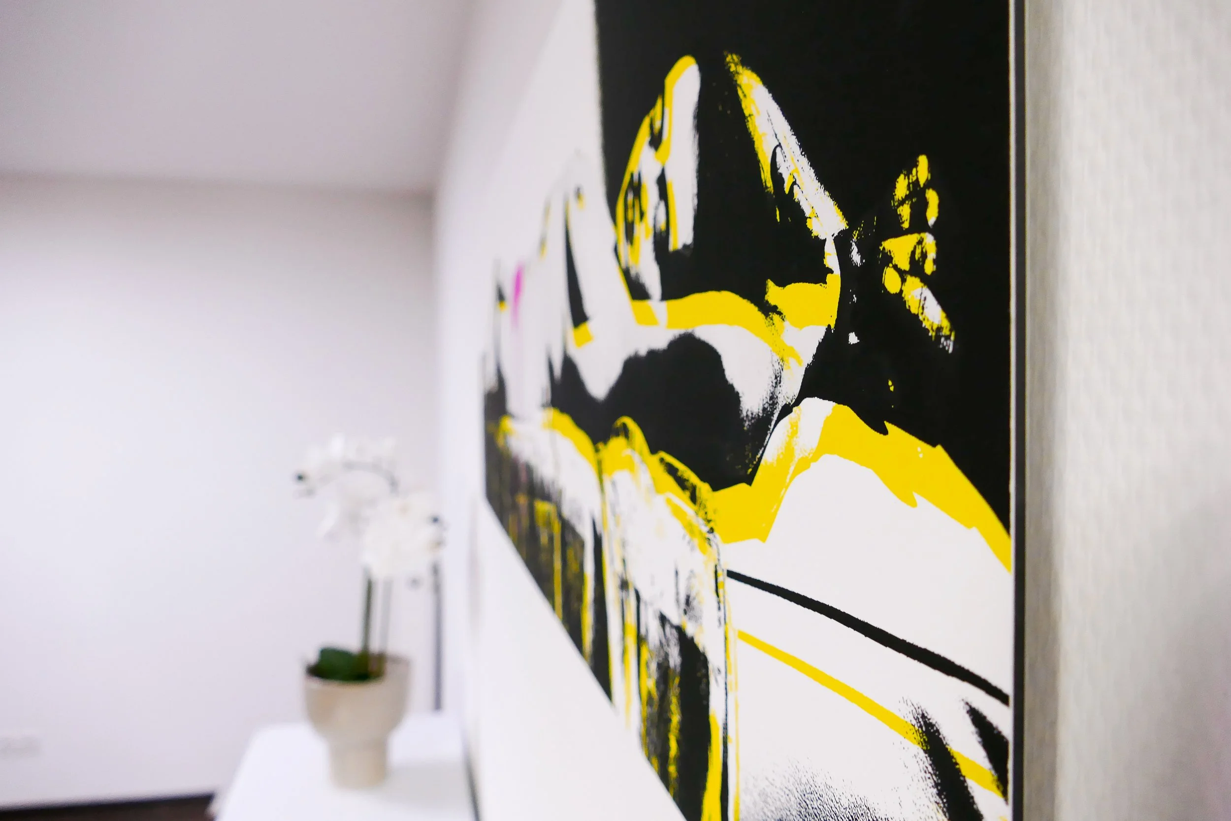 Nahaufnahme eines modernen, abstrakten Gemäldes mit schwarzer, weißer und gelber Farbgestaltung, an der Wand in einem Raum mit weißem Möbel und Pflanze im Hintergrund.