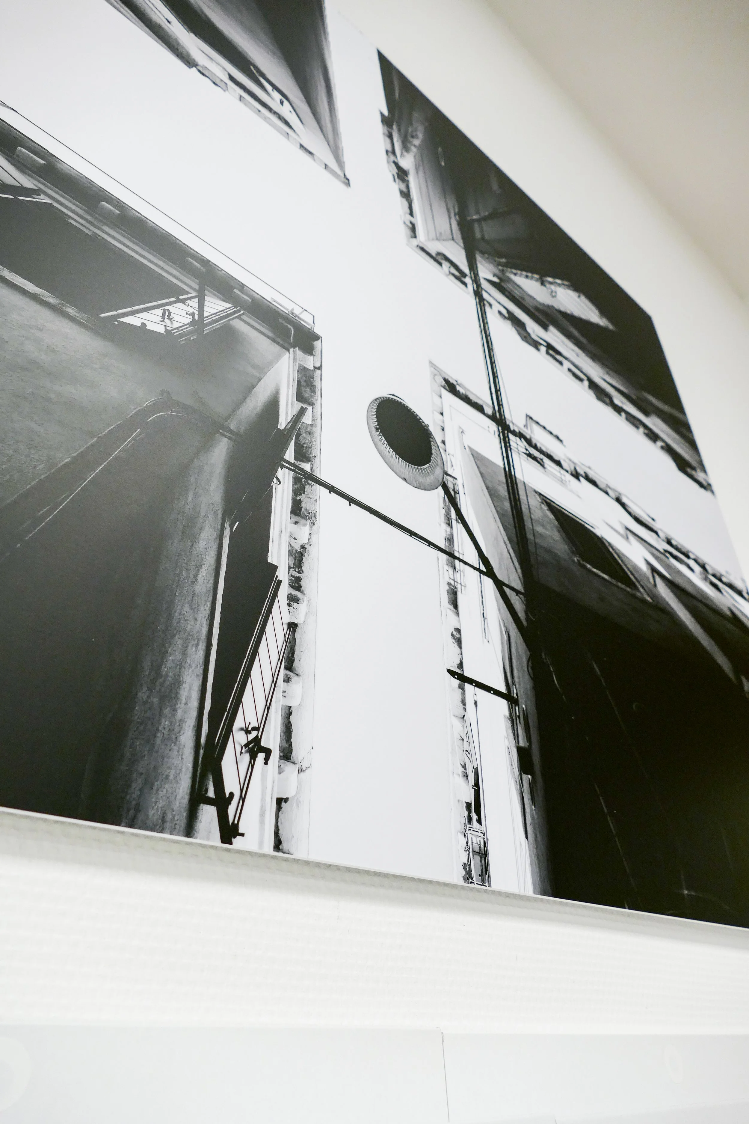 Schwarz-Weiß-Fotografie, die den Blick nach oben auf mehrere Gebäude zeigen, mit Balkon und einem runden Gegenstand, möglicherweise ein Lautsprecher oder ein Ventilator, an einem Kabel. Das Bild ist aus einer niedrigen Perspektive aufgenommen.