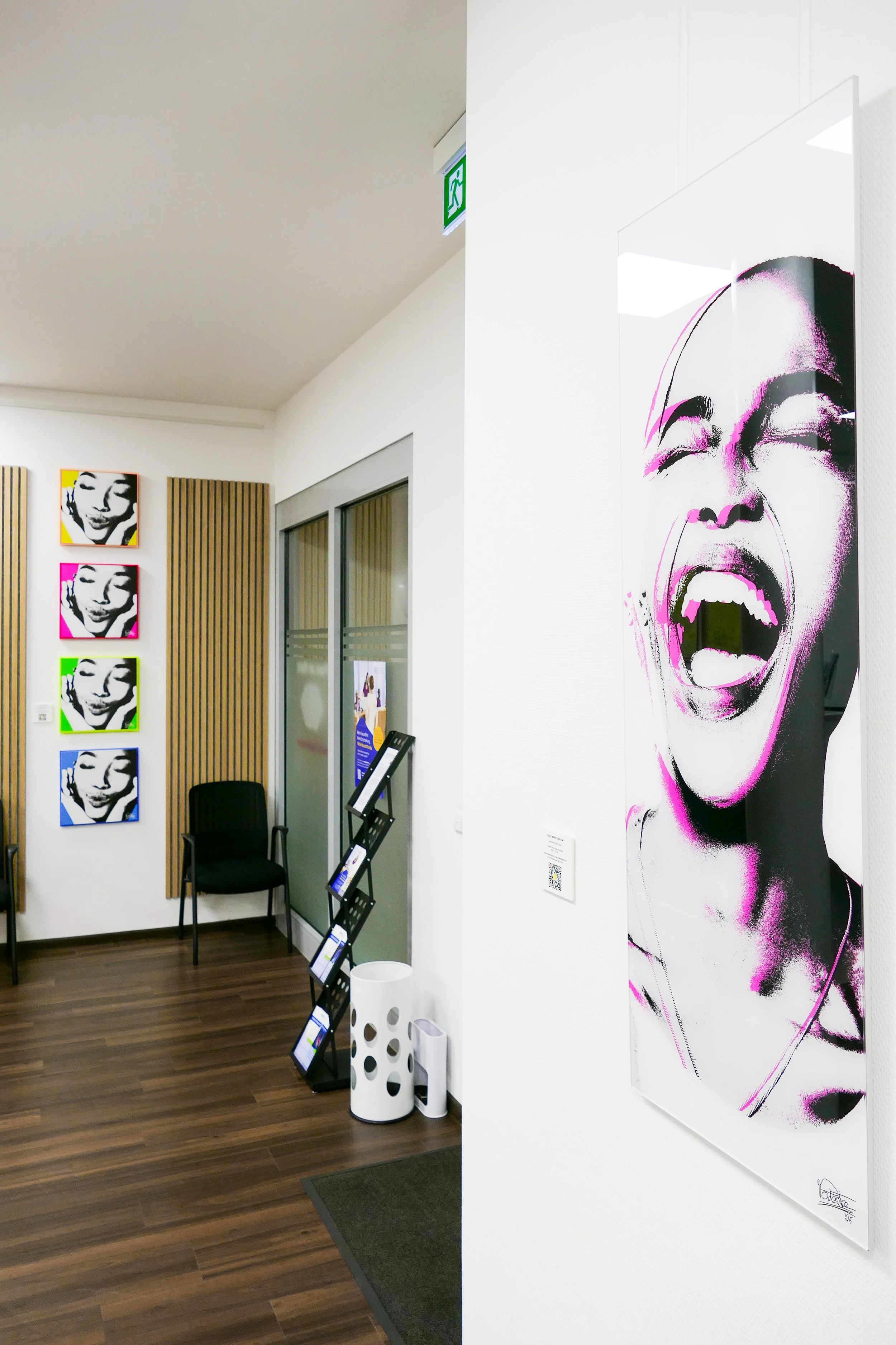Kunstwerke in einer Galerie, darunter eine große Schwarz-Weiß-Grafik eines lachenden Gesichts mit rosa Akzenten auf der rechten Seite. Auf der linken Seite sind vier farbige Portraits einer Person, die lächelnd und mit geschlossenen Augen, in gelb, p
