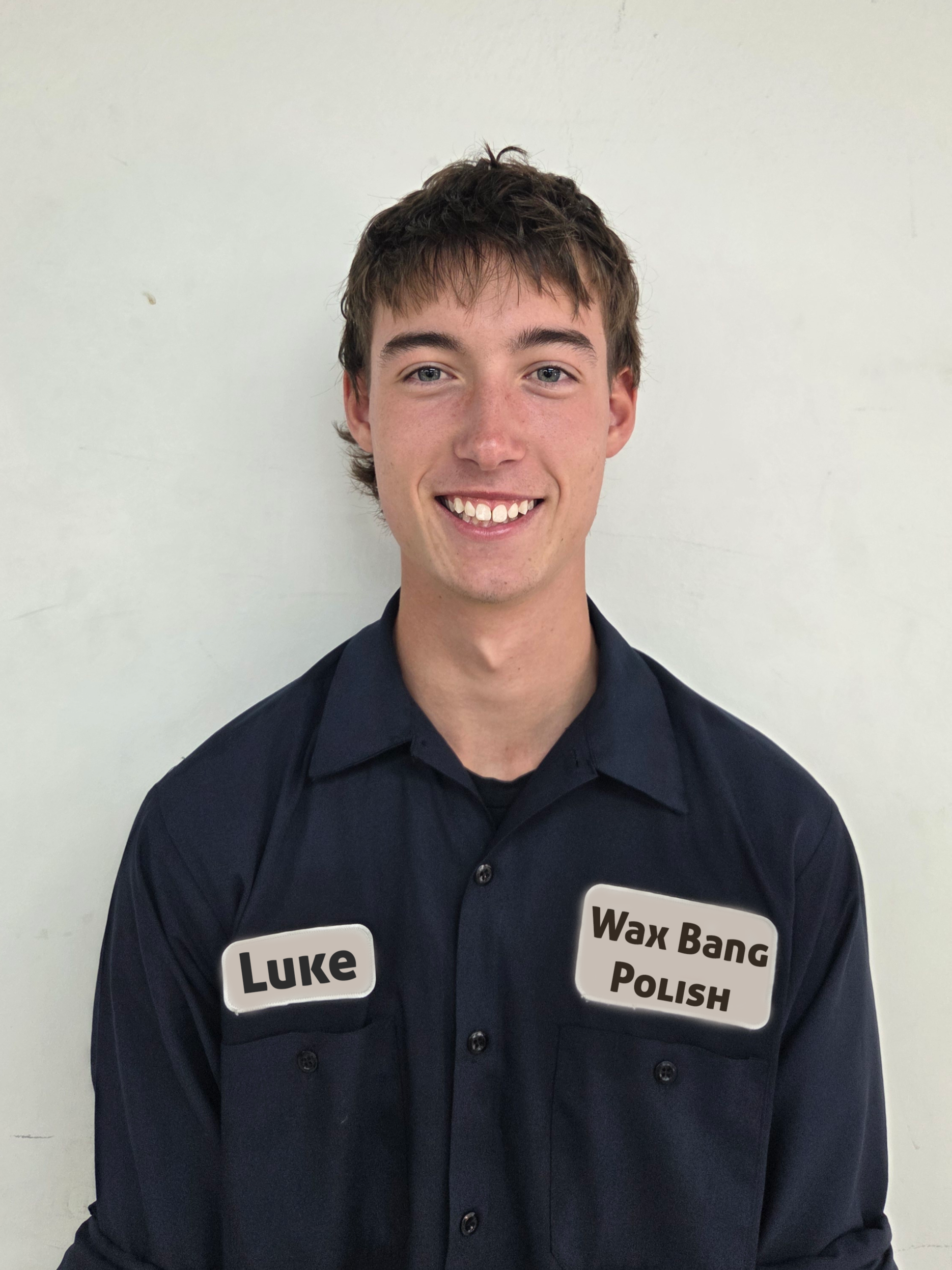 Young man with short brown hair smiling, wearing a dark uniform shirt with two name tags that read 'Luke' and 'Wax Bang Polish', standing against a plain white wall.