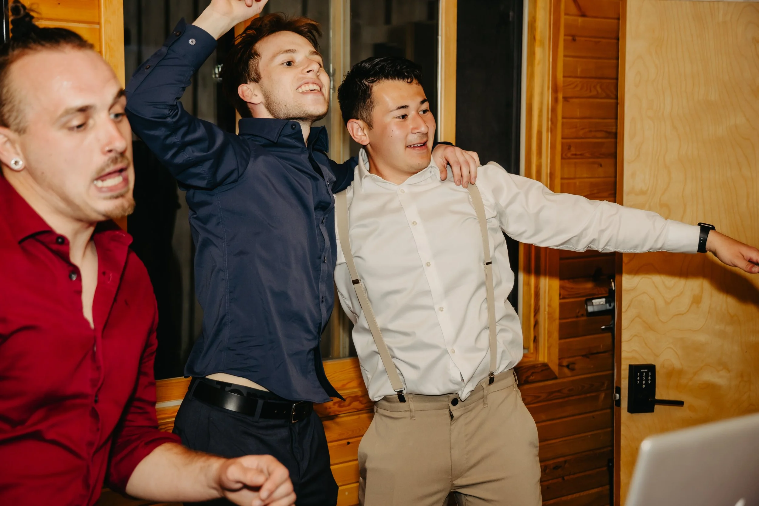 Group of three men celebrating, with one man raising his arm in excitement, inside a room with wooden paneling.