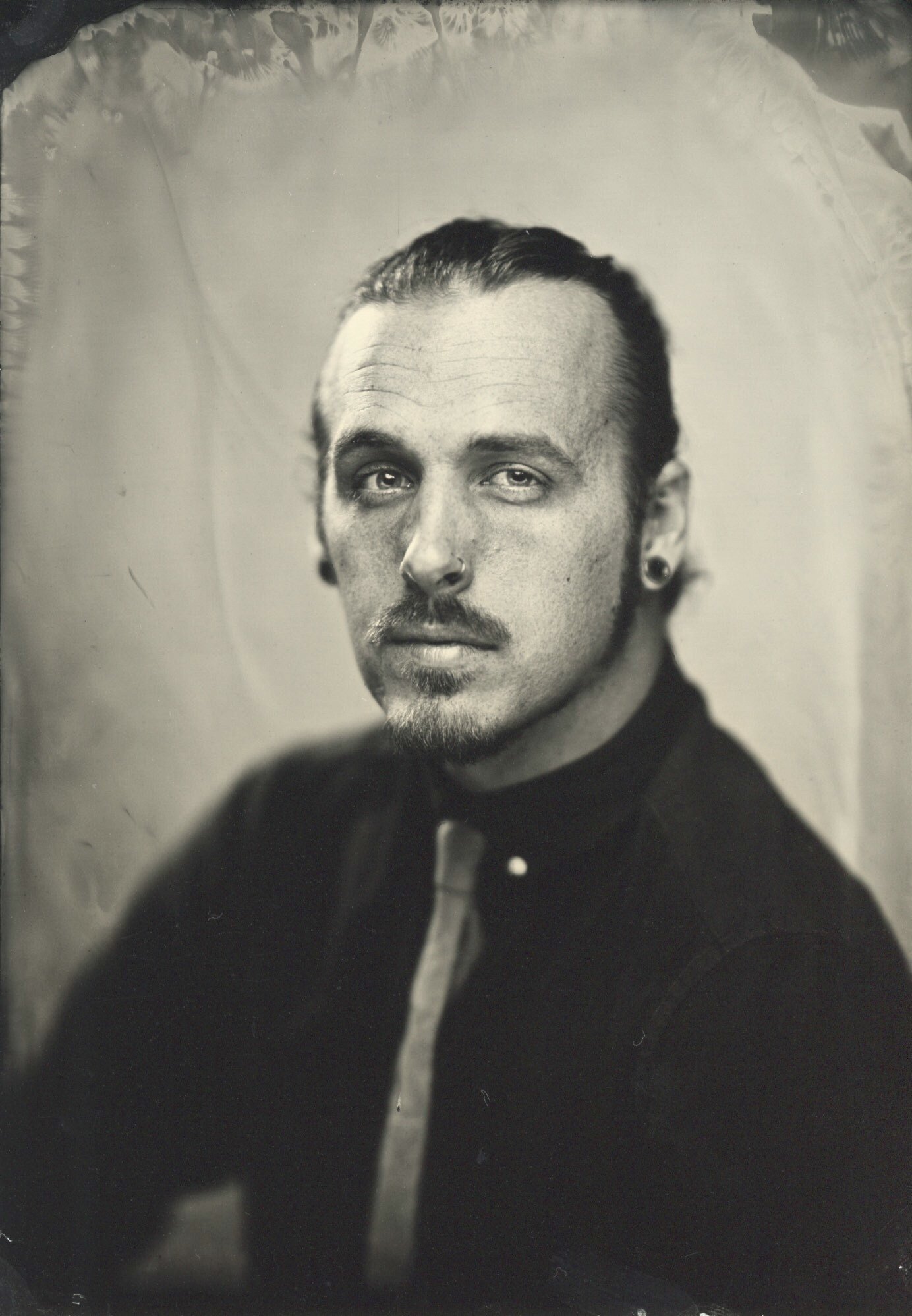 Black and white portrait of a man with slicked-back hair, facial hair, and multiple earrings, wearing a dark jacket.