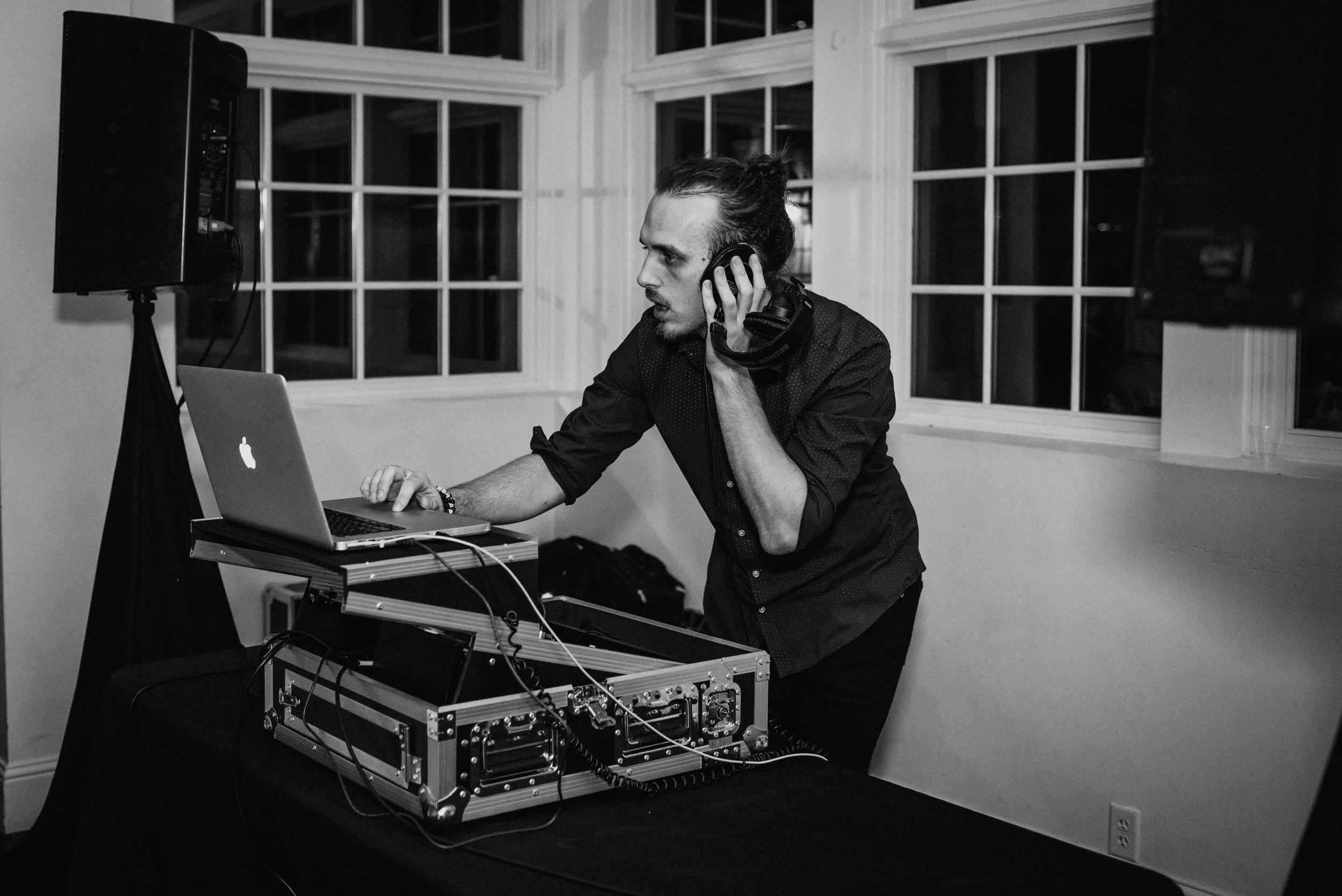 A man with long hair tied back, wearing a dark shirt, wearing headphones, operating electronic music equipment, with a laptop in front of him, in a room with large windows, black speakers, and various cables.