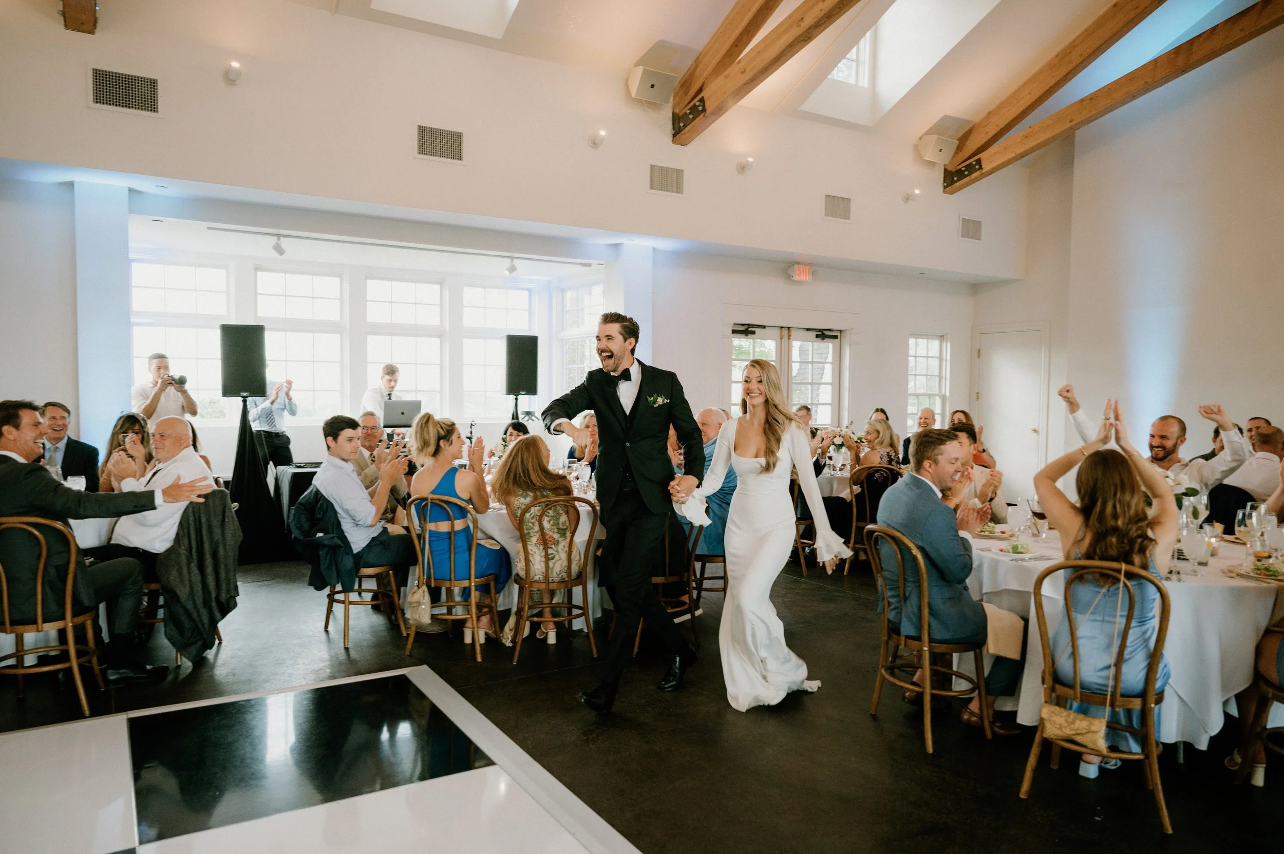 Bride and groom holding hands and smiling at their wedding reception with guests seated at decorated tables.